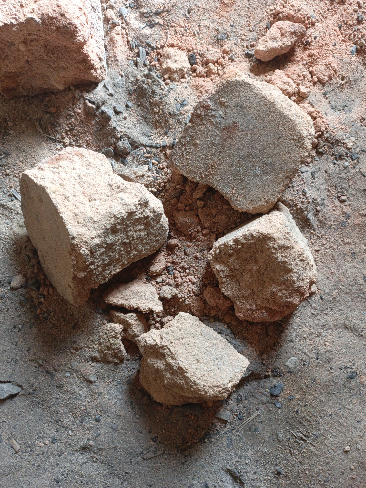 The image shows a close-up of scattered brick fragments on a surface composed of mixed-color earth and fine debris. Larger brick pieces are clustered in the center of the frame, exhibiting varied textures and shapes indicative of breakage. Smaller debris, including what appears to be pulverized brick and other particulate matter, covers the interstitial spaces and surrounding surface. The overall scene suggests a site where brick material has been broken down or degraded, potentially during or after a construction or demolition event. The location in the city of Unknown, Unknown, is speculative due to the lack of context or distinguishing features within the image.