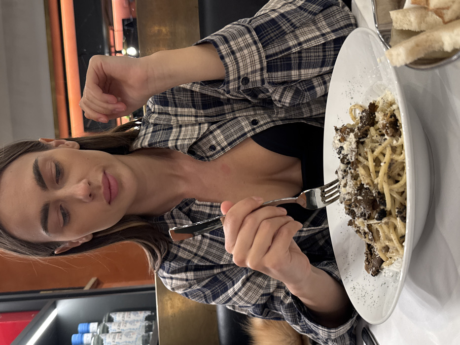 A young adult female is seated at a table within a restaurant in Verona, Italy, engaged in eating a meal. She is viewed from the chest up, with her gaze directed downwards towards a large white plate of spaghetti covered with dark truffle pieces and grated white cheese. Her right hand holds a silver fork, positioned over the pasta, with a matching knife resting next to it. She has long, dark brown hair and is wearing a blue and white plaid button-up shirt unbuttoned over a black low-cut top. The table is covered with a white tablecloth. To the lower right, a metal bowl contains several pieces of sliced white bread. The background features warm, possibly orange-toned walls, dark seating, and a shelf on the left displaying bottles of Acqua Panna mineral water, illuminated by linear lighting elements.
