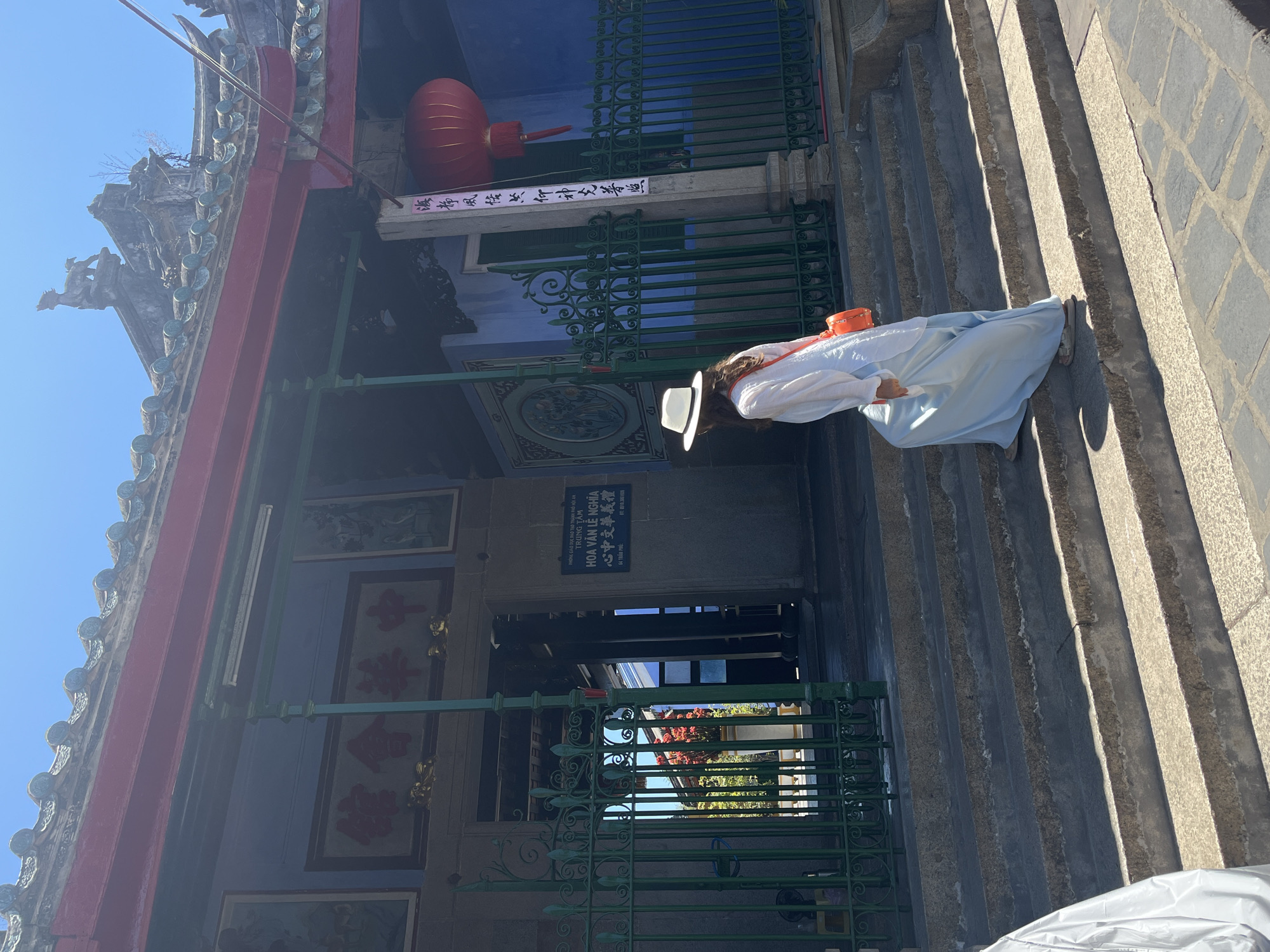 A person is depicted approaching the entrance of a traditional Asian building, likely a temple or cultural center, under bright daylight. The individual, seen from behind, wears a white long-sleeved top, a light blue flowing skirt or wide pants, and a white wide-brimmed hat, with an orange shoulder bag. They are ascending a set of rough-hewn stone steps towards ornate green metal gates that feature intricate scrollwork patterns.
The building itself exhibits traditional architecture, with a deep blue facade and red trim. A large, red cylindrical lantern hangs prominently above the entrance gates. The roof boasts decorative grey tiles and intricate sculptural elements, including what appears to be a mythical creature carving on a corner.
Several signs with text are visible. Directly above the gates, a rectangular sign in Vietnamese reads: "PHÒNG GIÁO DỤC ĐÀO TẠO THÀNH PHỐ HỘI AN TRUNG TÂM HOA VĂN LÊ NGHĨA" (Hoi An City Education and Training Department - Le Nghia Chinese Cultural Center). Beneath this, Chinese characters read "心中文華義禮" (Xin Zhong Wen Hua Yi Li), followed by the address "64 TRẤN PHỦ" and a phone number (0511.386195). Another partially visible sign to the left of the entrance displays the Chinese characters "館會" (Guǎnhuì), suggesting a guild or assembly hall. The scene is set on a clear, sunny day, in Hội An Ward, Vietnam, showcasing its rich cultural heritage.