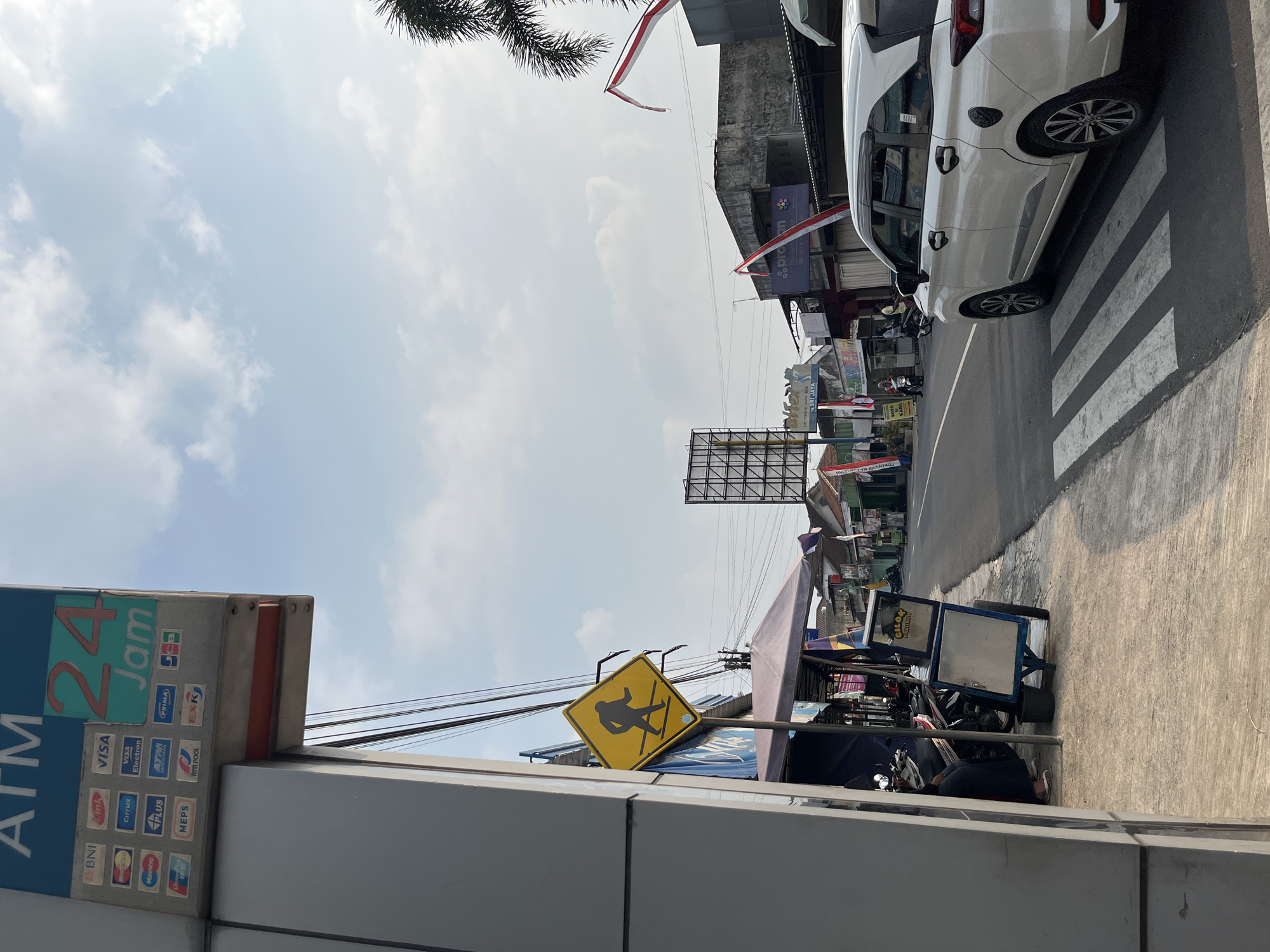 The image captures a street scene in Matesih, Indonesia, under a partly cloudy sky. In the foreground, a white car is stopped at a pedestrian crossing. To the left, part of an ATM enclosure is visible, displaying logos of various banks and credit card companies, along with "ATM 24 Jam". Further down the street, small businesses with awnings line the road, and several motorcycles are parked. A yellow diamond-shaped traffic sign with a pedestrian icon is mounted on a pole. Red and white Indonesian flags are displayed on some buildings. In the distance, a large billboard stands against the sky. The overall environment suggests a typical urban street in Indonesia.