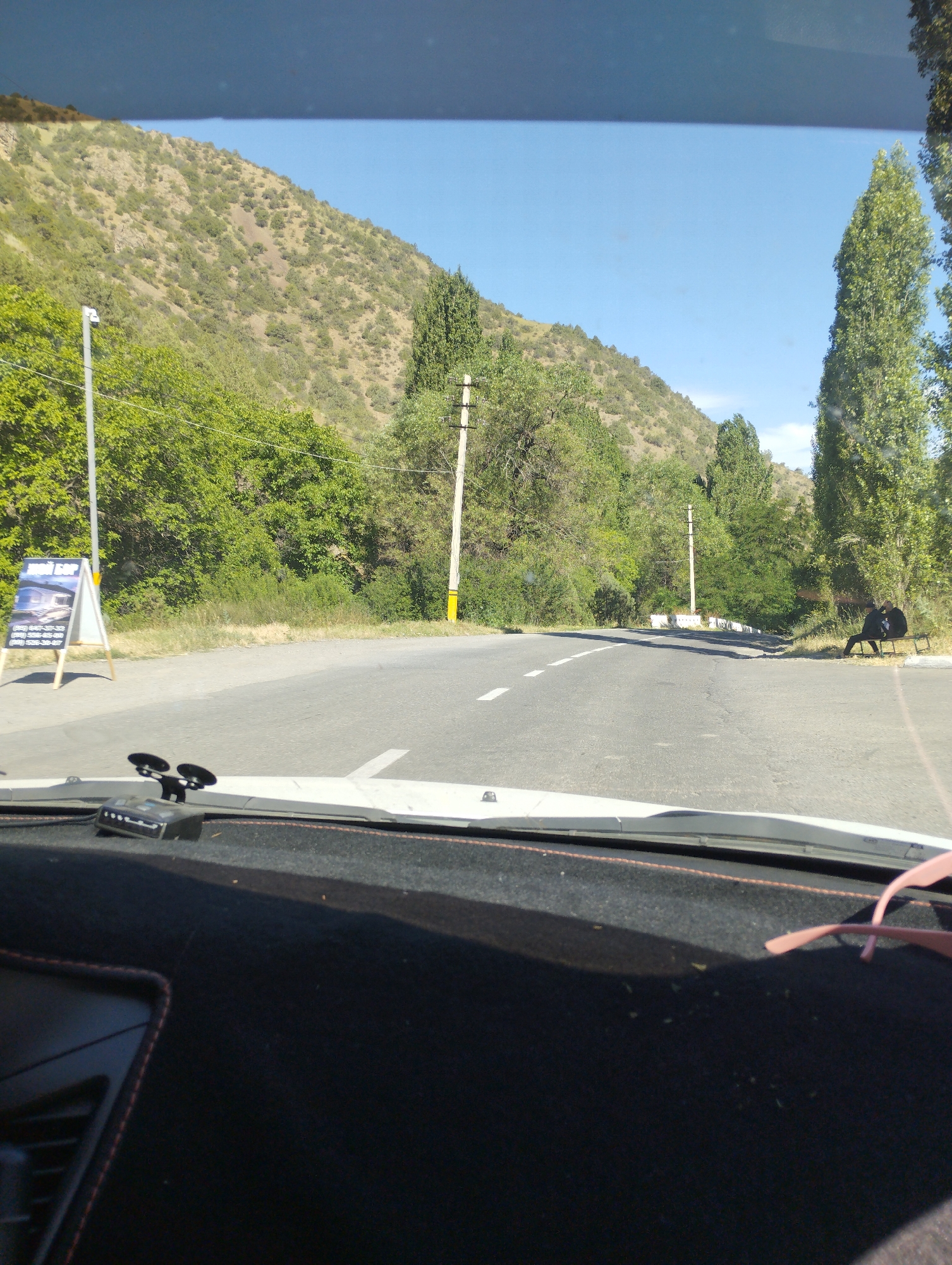 The image, taken from inside a vehicle, depicts a road curving through a mountainous and tree-lined landscape. On the left side of the road, a white A-frame sign with images and text is visible. The road itself is asphalt with white dashed lines indicating lanes. In the distance, two individuals are seated on a bench on the right side of the road. Tall green trees line both sides of the road, with a barren, rocky hillside forming the backdrop. The sky is clear and blue. The vehicle's dashboard is in the foreground.