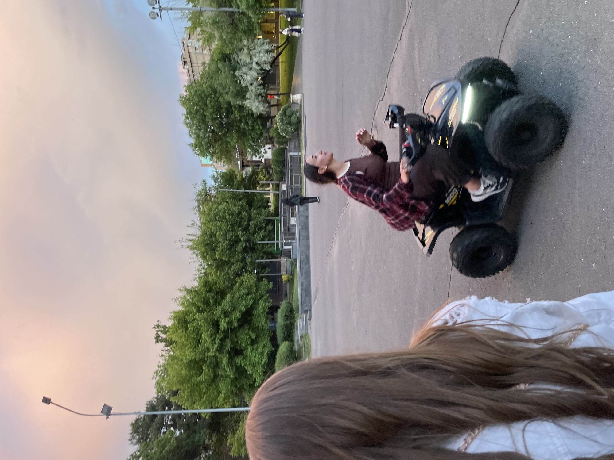 A woman is riding a small, black and yellow ATV on a paved area, likely a sidewalk or plaza. She is wearing a maroon and black plaid shirt over a brown top, with black pants and white sneakers. Her head is tilted back. Trees are in the background, and a street lamp is visible on the left. Another person, with long brown hair and wearing a white shirt, is partially visible in the lower portion of the frame.
While Yerevan, Armenia is known to have a considerable amount of greenery, the architecture is not visible, so location cannot be confirmed by elements present in the image.
