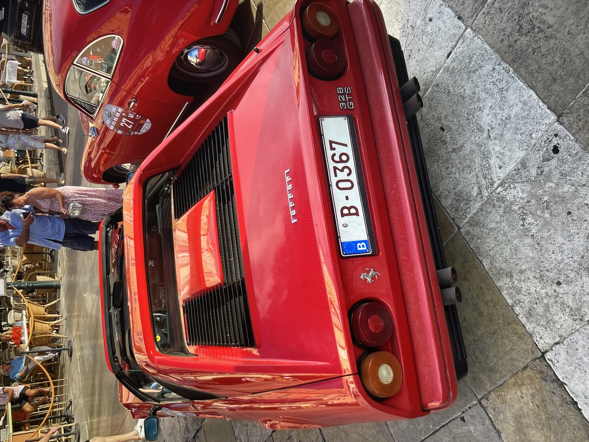 The image captures the rear of a red Ferrari 328 GTS, license plate B-0367, parked on a stone-paved street in a city setting. The photograph is taken at a slight angle, showing the car's rear grills, taillights, and exhaust pipes. Partially visible in the upper left is another vintage red car, with people around it. The surroundings suggest a public space, possibly a square or street with outdoor seating, given the presence of tables and chairs in the background. People are casually dressed and are standing or sitting, implying a relaxed atmosphere. The specific location within the city of Unknown, Unknown is not identifiable from the image.