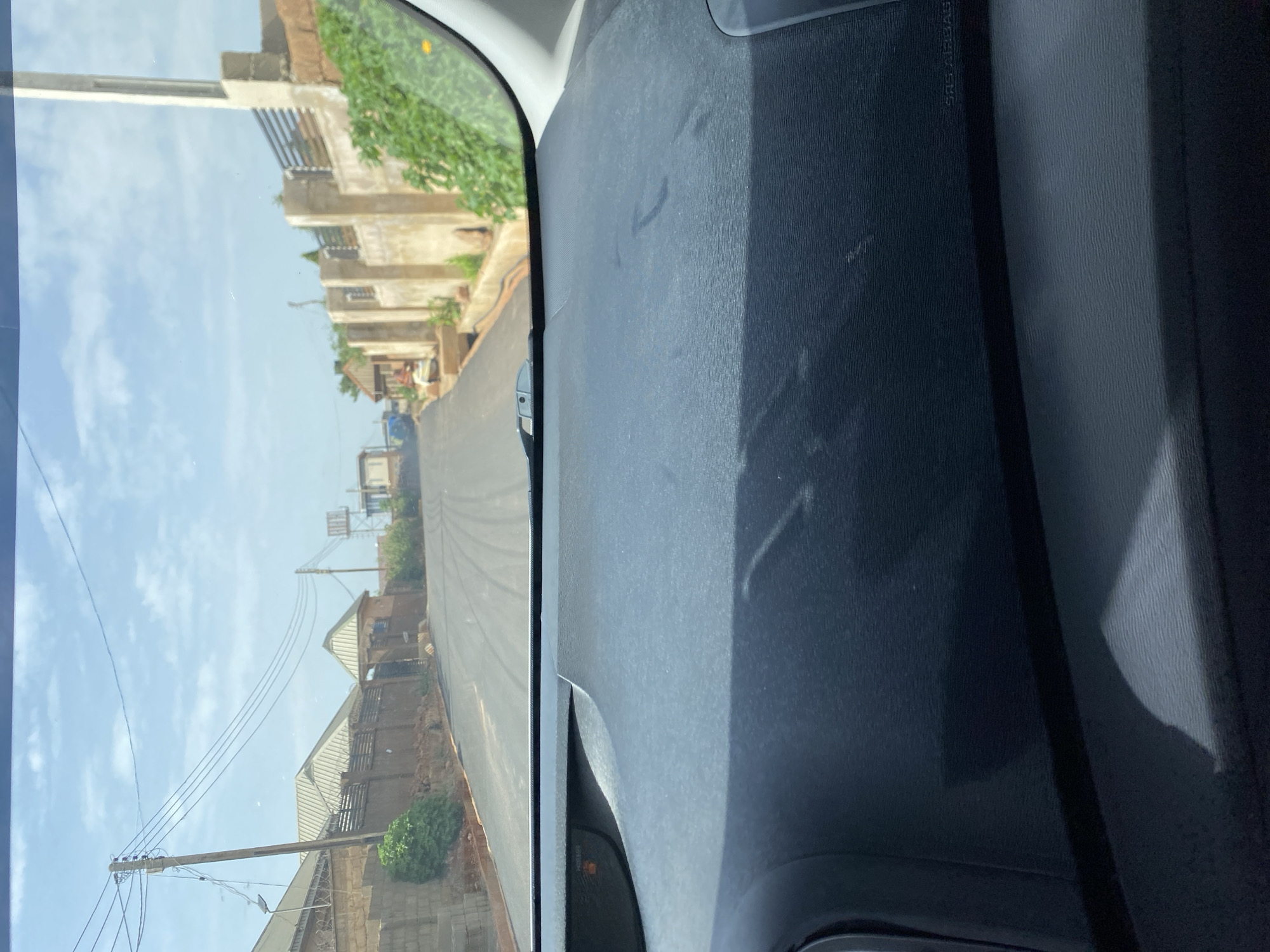 This image is a photograph taken from inside a vehicle, looking out through the windshield. The view is of a paved road winding uphill through a residential area. On either side of the road, there are houses with brick walls and metal roofs, some with fences. There are also utility poles with overhead power lines. The sky is partly cloudy. The dashboard of the vehicle is visible in the foreground. The location is identified as Bar Jirgi Summa, Nigeria.