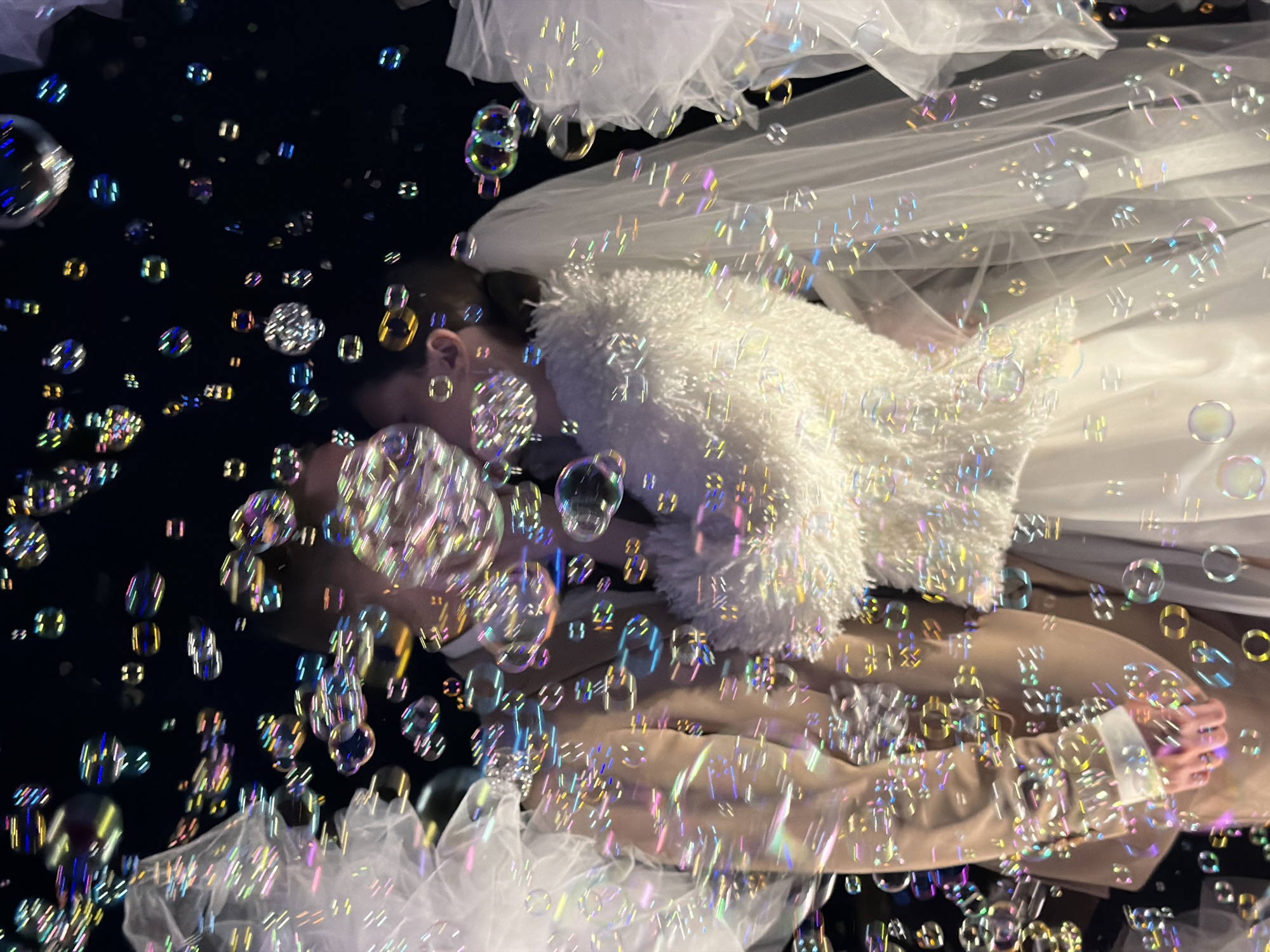 A couple is seen from an elevated perspective, presumably kissing behind a dense layer of soap bubbles. The female figure is wearing a white fuzzy shawl and a semi-transparent veil. The male figure is wearing a tan suit. The background has both light and dark sections, some of which are obscured by the bubbles. There is no definitive evidence of location. Therefore, the city of Chaikovsky, Russia cannot be identified.