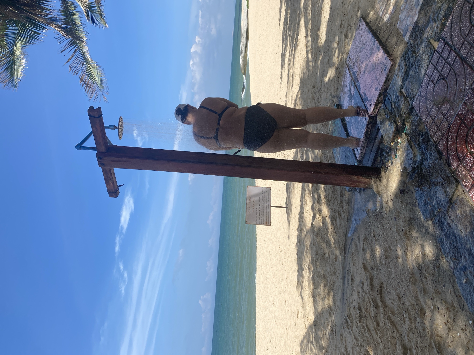 An outdoor beach shower on a sandy beach in Phu Thuy Ward, Vietnam, is depicted under a clear blue sky with sparse white clouds. A solitary individual, viewed from the back, stands under the active shower, wearing a black two-piece swimsuit and a light-colored cap or towel on their head. Water streams from a circular, brass-colored shower head mounted on a rustic wooden structure. Below the person, several wet, flat stepping stones are positioned on the sand, with water pooling and flowing towards patterned red tiles in the foreground. The wide, light-colored sandy beach extends towards the tranquil, light blue-green sea, where gentle waves are visible near the shore. A white warning sign, partially obscured, is planted in the sand to the left, displaying text in Vietnamese and English, cautioning against swimming due to "ROUGH SEA, HIGH WAVES." A palm tree's fronds are visible in the upper right, casting shadows on the sand.