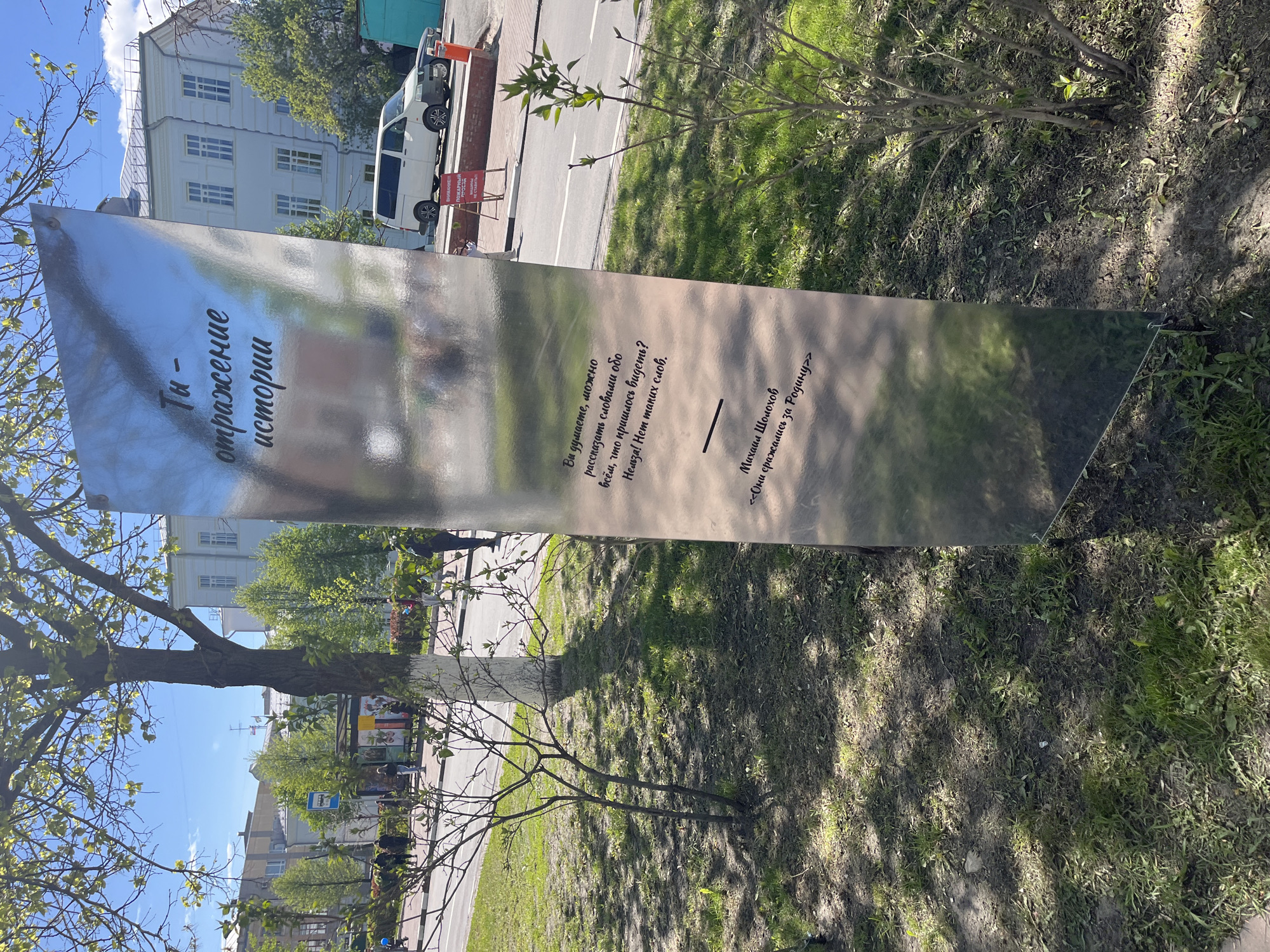 The image shows a shiny, reflective metal sign erected outdoors, apparently in a park in the city of Ulyanovsk, Russia. The sign features text in Russian, and reflects the surrounding urban environment, including buildings, trees, and the sky. The sign is rectangular, angled, and stands on the ground surrounded by grass and foliage. A road with parked vehicles is visible in the background.