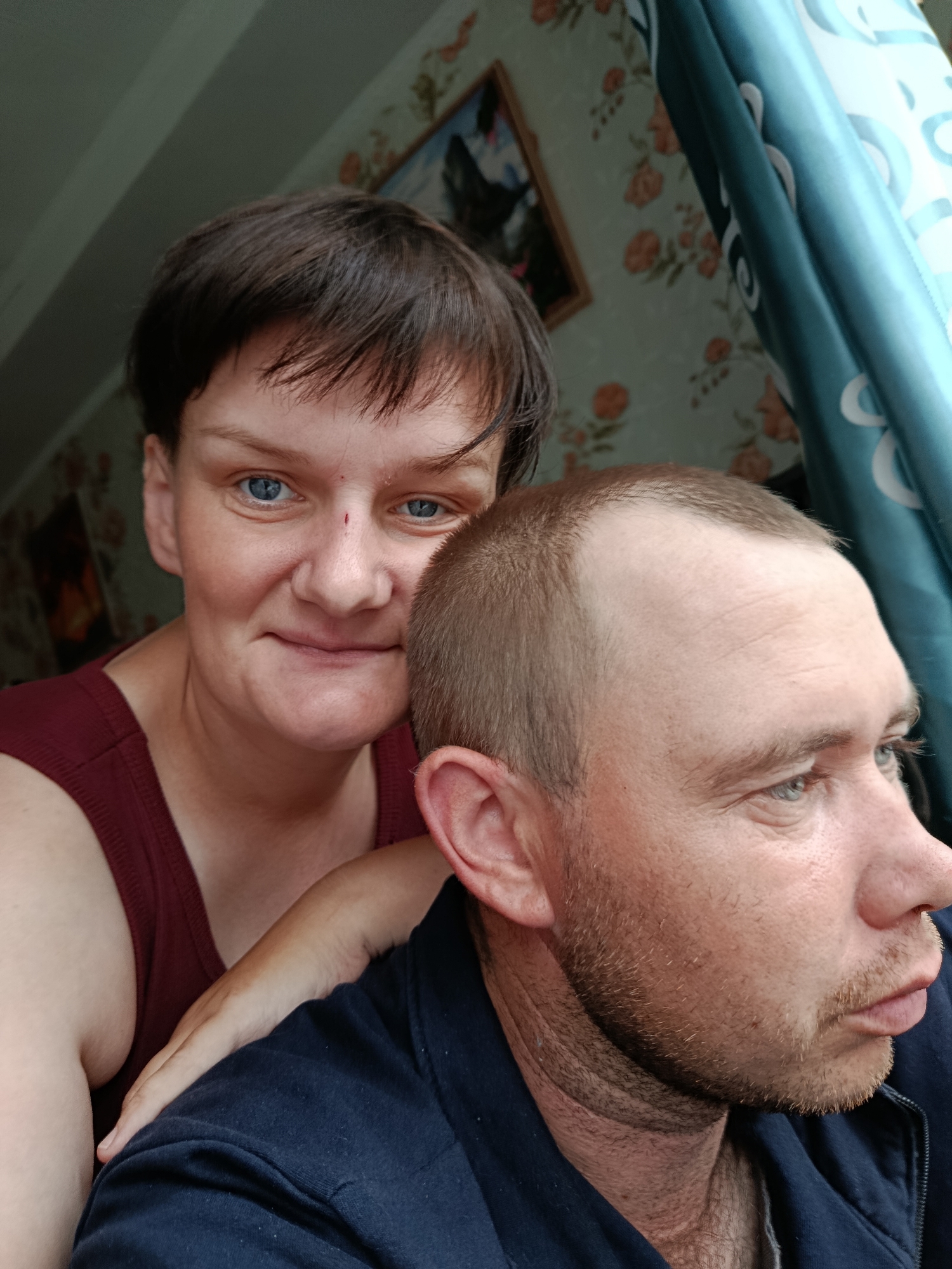 This media file features a close-up, selfie-style shot of a man and a woman. The woman is in the foreground, her head and shoulders are visible, and she is looking directly at the camera with a slight smile. She has short, dark hair, blue eyes, and fair skin. A small red mark is visible on the bridge of her nose. She is wearing a dark red sleeveless top.
Behind and to the right of the woman, the man's profile is visible. He has short, light brown hair, and is looking to the right of the frame. He has light blue eyes and a light beard. He is wearing a dark blue zippered jacket over a grey t-shirt. The woman's arm is resting on his shoulder.
The background suggests an indoor setting. The wall behind them is covered with floral wallpaper in shades of green and peach. To the right, a blue curtain with a circular pattern is visible. The lighting suggests it is daytime, possibly indoors with natural light from a window. The overall mood appears relaxed and intimate. The image was taken in Foky, Russia. No visible text is present in the media file.
