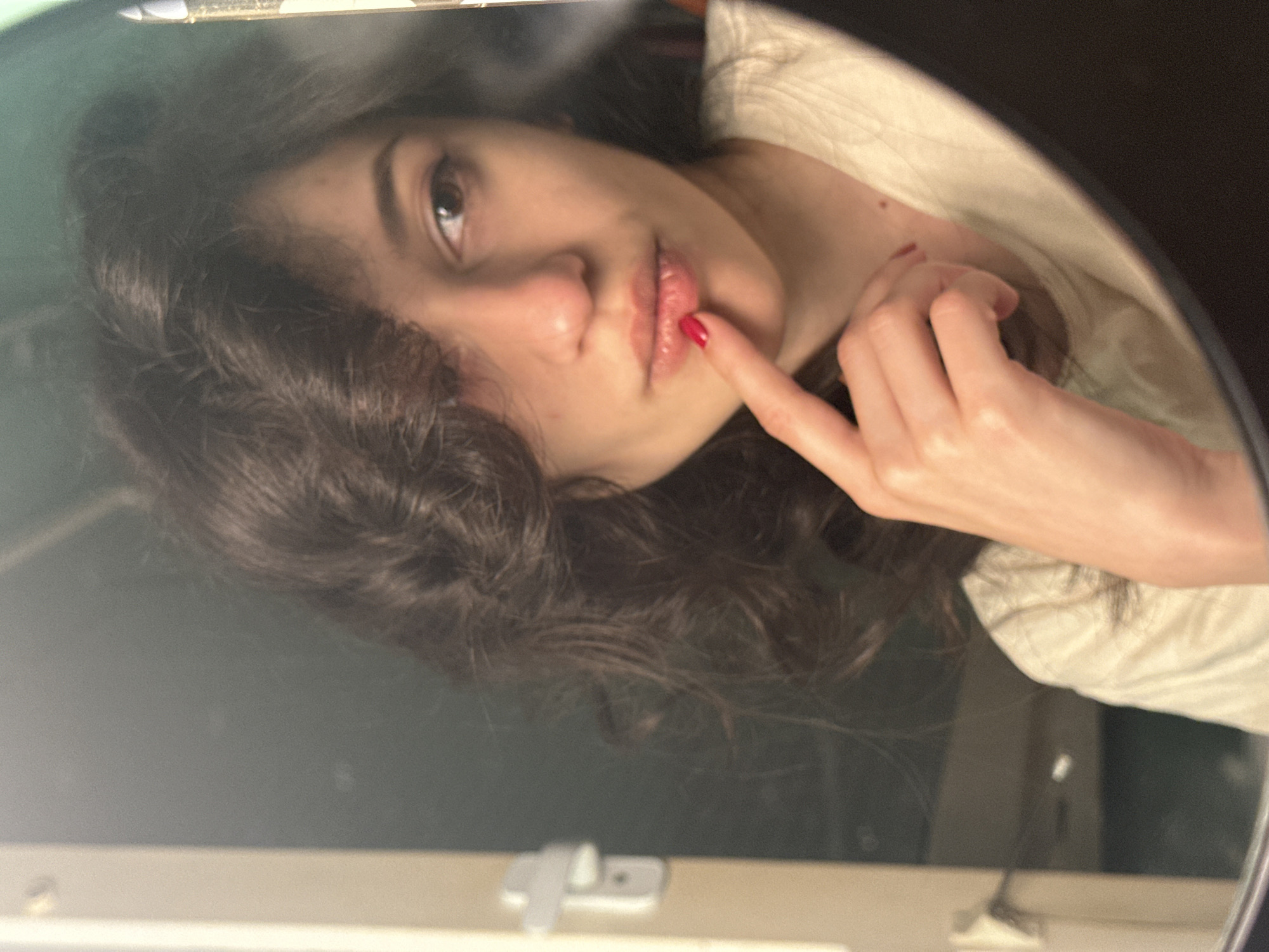 A young woman with dark, wavy hair is seen in a reflection, likely in a mirror. She is wearing a light-colored t-shirt and has her right index finger touching her lips. Her lips are painted with red lipstick, and her fingernail on that finger is also painted red. She has brown eyes and a light complexion. The background appears to be a room with a doorframe visible on the left, suggesting an indoor setting. The lighting is somewhat dim, creating soft shadows. There is no information to suggest this image is located in Sofia, Bulgaria.