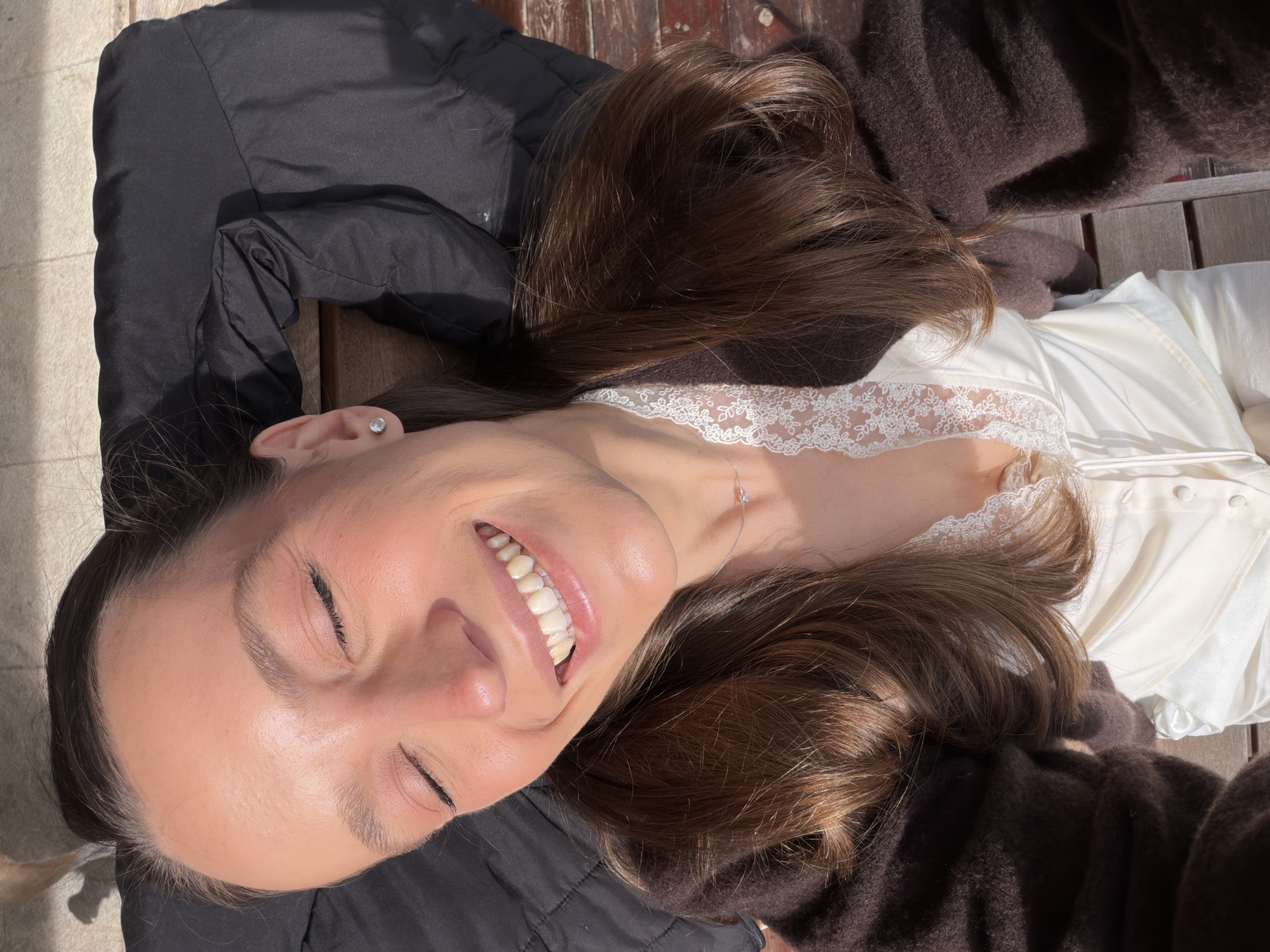 A woman with brown hair is lying down, smiling with her eyes closed. She is wearing a white top with lace detailing around the neckline. She is also wearing a black jacket and a brown sweater. The background appears to be wooden planks. The location in the city of Trebesin, Montenegro is not discernible from the image.