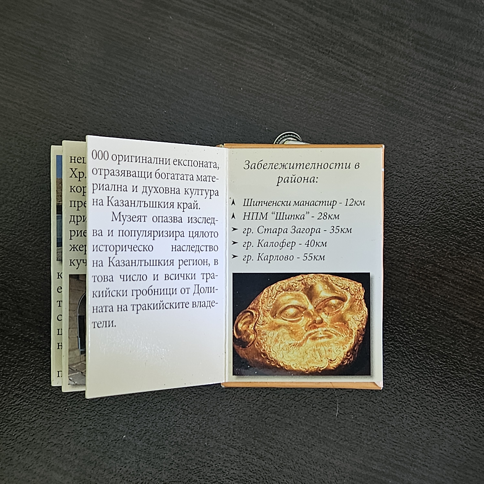 The image shows a small, open booklet lying on a textured surface. The booklet appears to be a souvenir or guide, possibly related to the city of Stara Zagora, Bulgaria. One open page lists "Sights in the Region" with distances to several locations, including the Shipka Monastery (12km), the Shipka National Park Museum (28km), Stara Zagora (35km), Kalofer (40km), and Karlovo (55km). Below the list is a photograph of a gold artifact, possibly a mask or face. The opposite page contains text in Bulgarian, discussing artifacts, cultural heritage, and Thracian tombs in the Kazanlak region. Other pages are visible to the left, with snippets of text and a possible image. The overall impression is that of a travel guide or informative brochure about historical sites and treasures in and around Stara Zagora, Bulgaria.