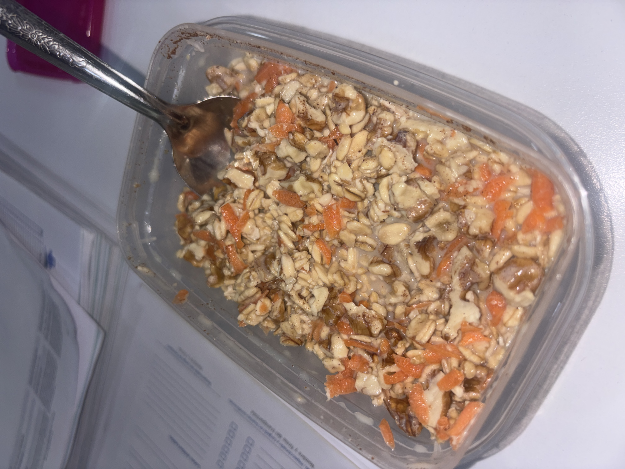 A plastic container of food sits on a white surface, partially obscuring stacks of papers. The food appears to be a mixture of oats, chopped carrots, and nuts in a liquid, possibly milk or yogurt. A spoon rests in the container, presumably for eating. The environment suggests an indoor setting, likely a desk or table due to the presence of paperwork. The location of Buenavista, Mexico, is not discernible from the image.