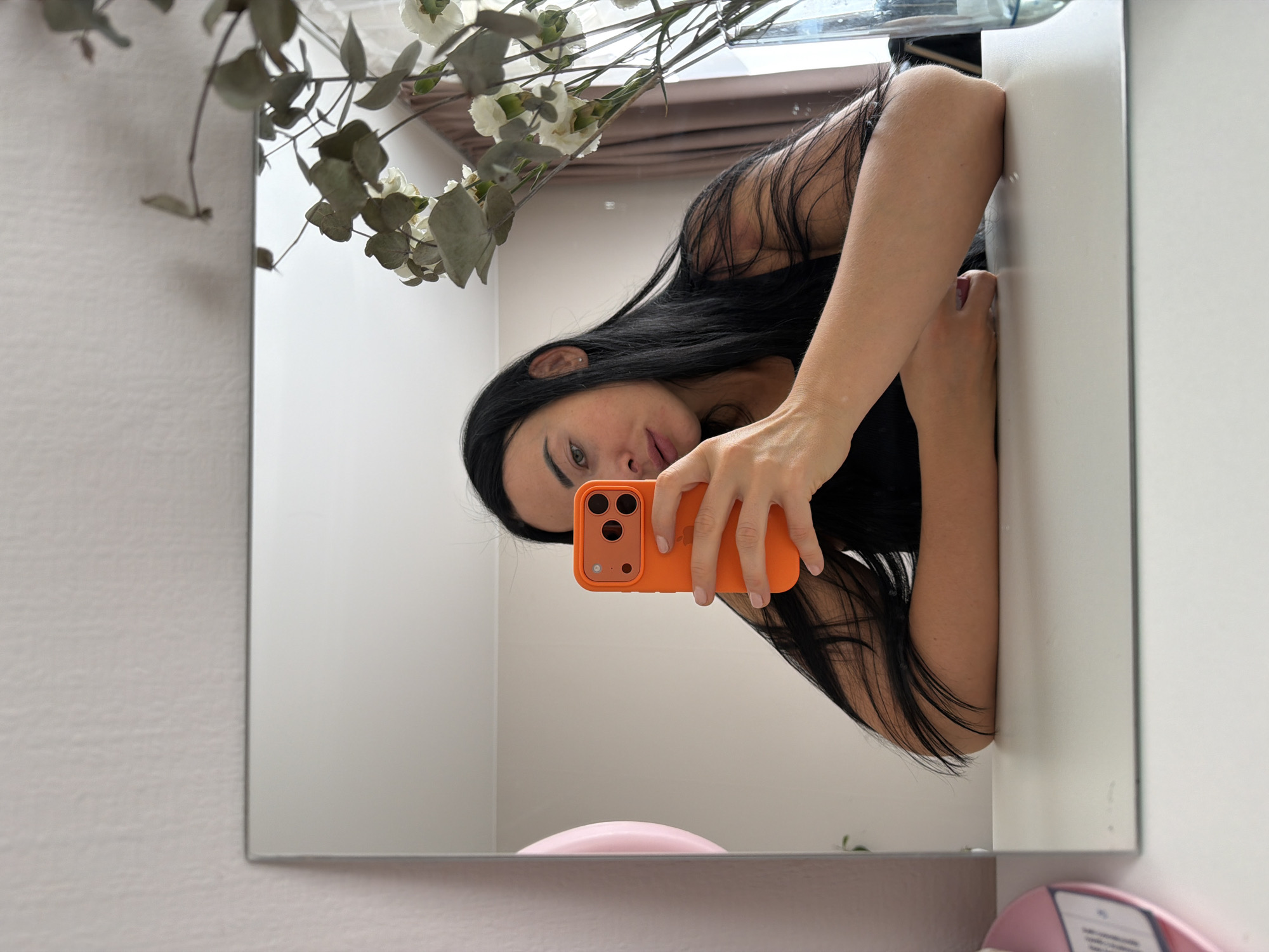 A woman with long, dark hair is taking a selfie in a mirror. She is holding an orange phone in her right hand. The mirror reflects her face, part of her upper body, and the surrounding environment. The background appears to be a room with white walls. There is a plant with white flowers and greenery in the upper left corner of the mirror's reflection. The location and details are insufficient to identify the scene as being in Saint Petersburg, Russia.