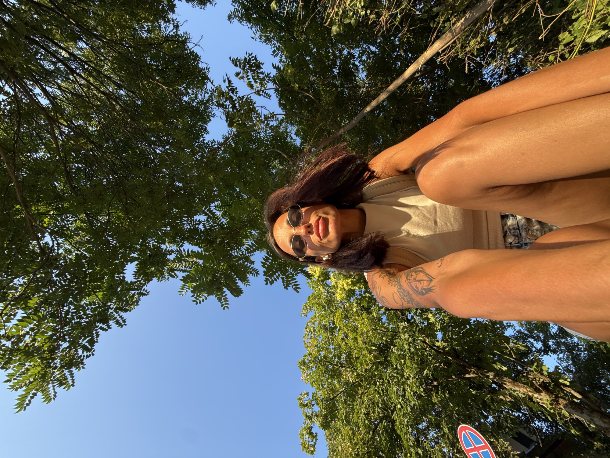 A low-angle, wide shot shows a woman sitting in an outdoor setting with trees and a blue sky overhead. The woman is wearing a tan tank top, denim shorts, and sunglasses. Her arms and legs are visible, and she has tattoos on her left arm. There is a red and blue sign visible on the left side of the image. The scene appears to be in the city of Pleven, Bulgaria, as indicated by the signage which bears Bulgarian text and symbols. The lighting suggests it is daytime.