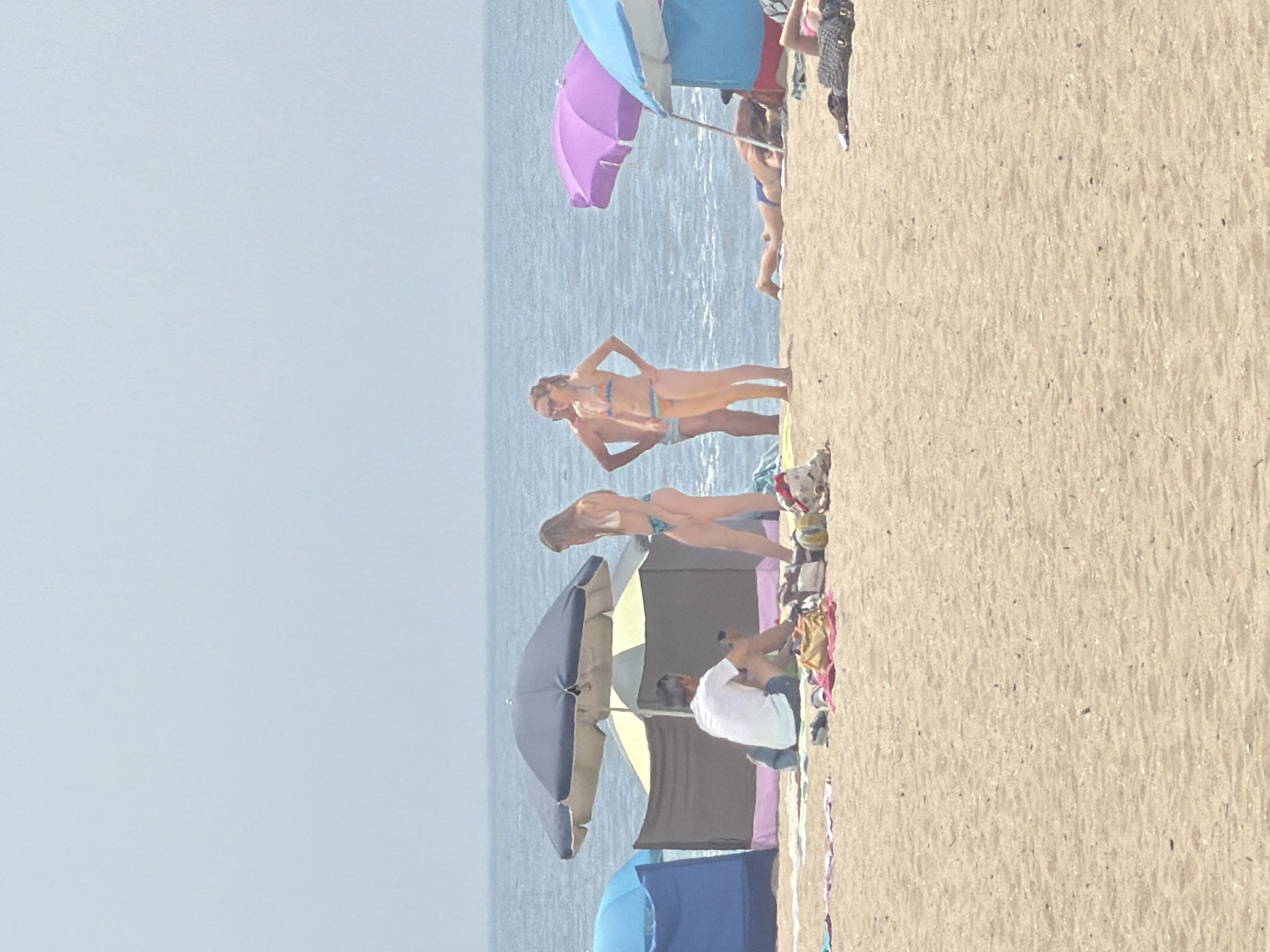 The image depicts a beach scene with several people enjoying a sunny day. In the foreground, on the sandy beach, a man wearing a white t-shirt and blue shorts is seated, facing away from the camera, appearing to be looking at his phone. Next to him are various beach items like towels and bags.
Further back, there are several people and beach umbrellas. Two women are standing near a beach tent and an umbrella. One woman is wearing a light-colored bikini, and the other is wearing a multicolored bikini. To their right, another woman is lying down on the sand, partially visible. Another individual is lying on their side further in the background. There are also several beach umbrellas and tents set up on the sand.
The ocean is visible in the background, with calm water and a hazy sky. The overall impression is of a leisurely day at a beach.