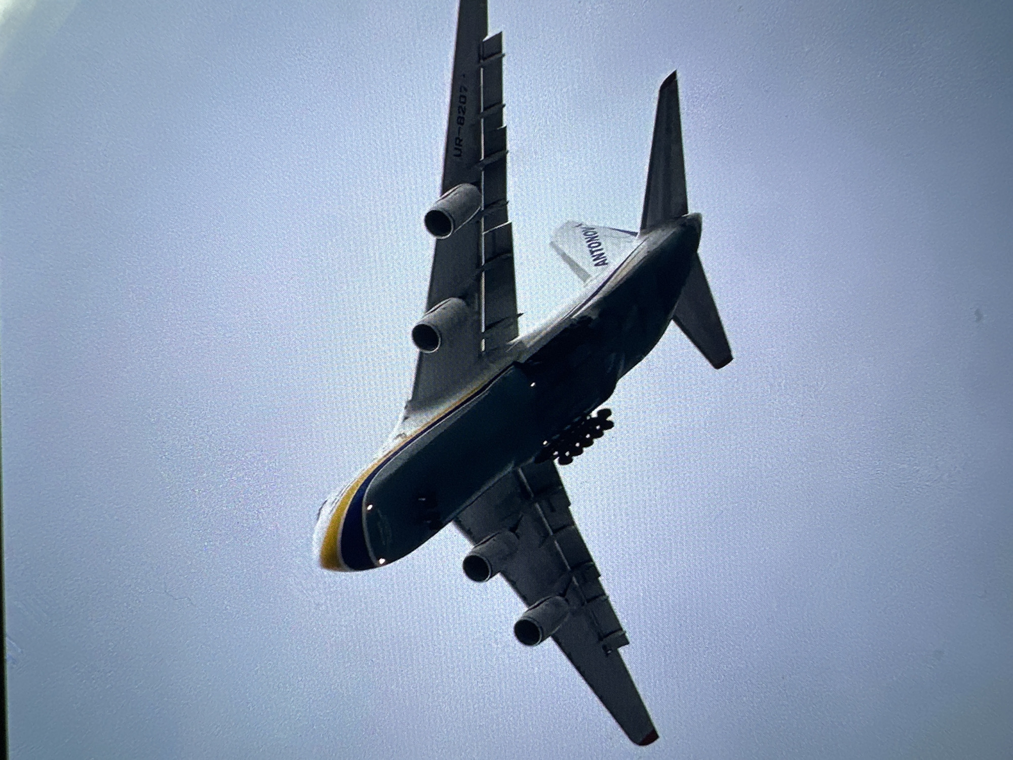 An airplane, specifically an Antonov AN-124, is flying. The aircraft is painted with a yellow and blue stripe near the front, and the words "ANTONOV" are visible on the tail. The registration number "UR-8207" is visible on the wing. The plane is against a grey sky. Location is Unknown, Unknown.