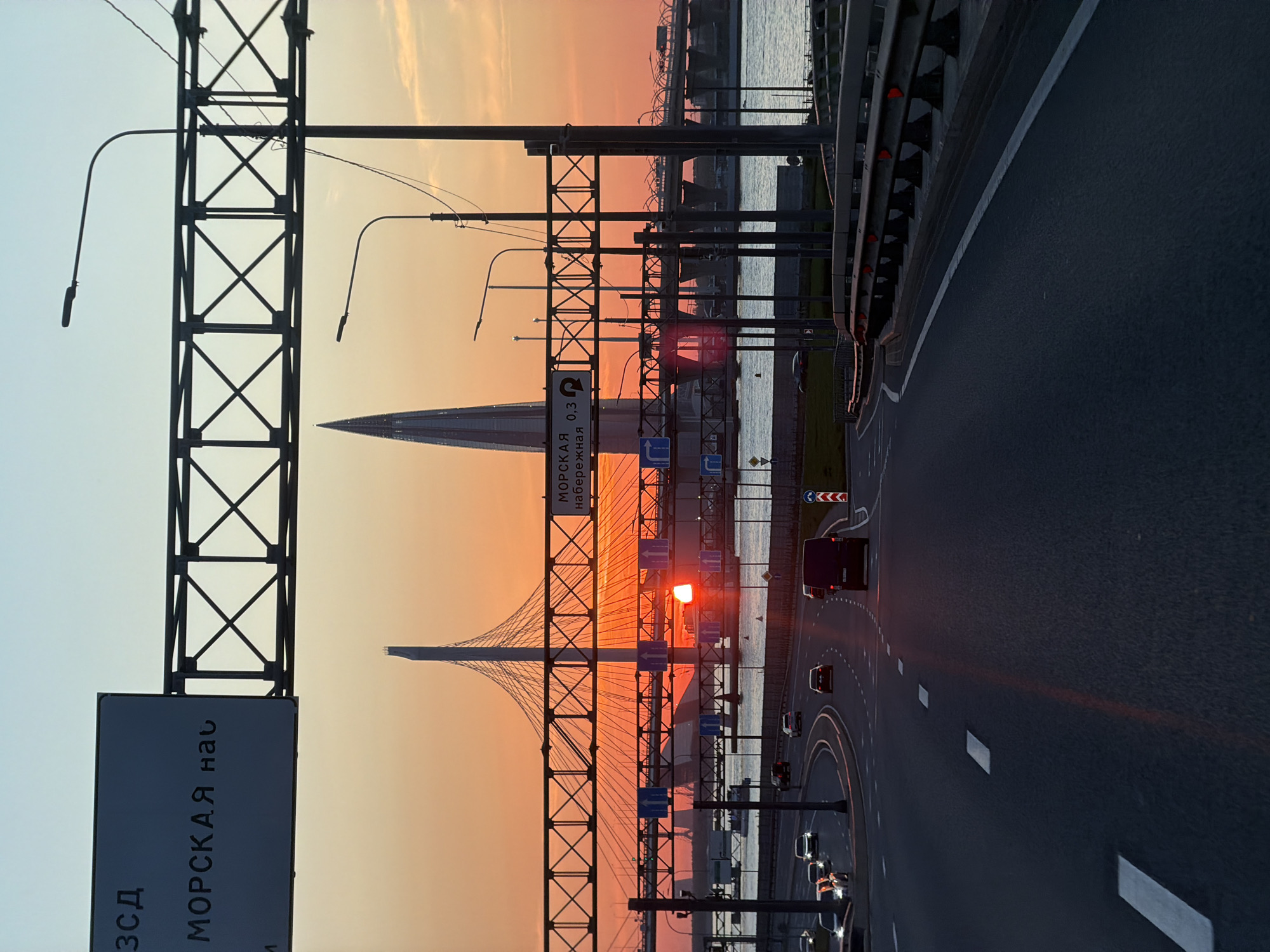 The image captures a sunset view in Saint Petersburg, Russia, featuring the Western High-Speed Diameter (ZSD) highway. The Lakhta Center skyscraper is visible in the background. The sky displays a gradient of colors from light blue to orange as the sun sets. A cable-stayed bridge spans the water, and the highway curves to the right, with several vehicles traveling on it. Metal road infrastructure, including support structures and signage, are in the foreground.