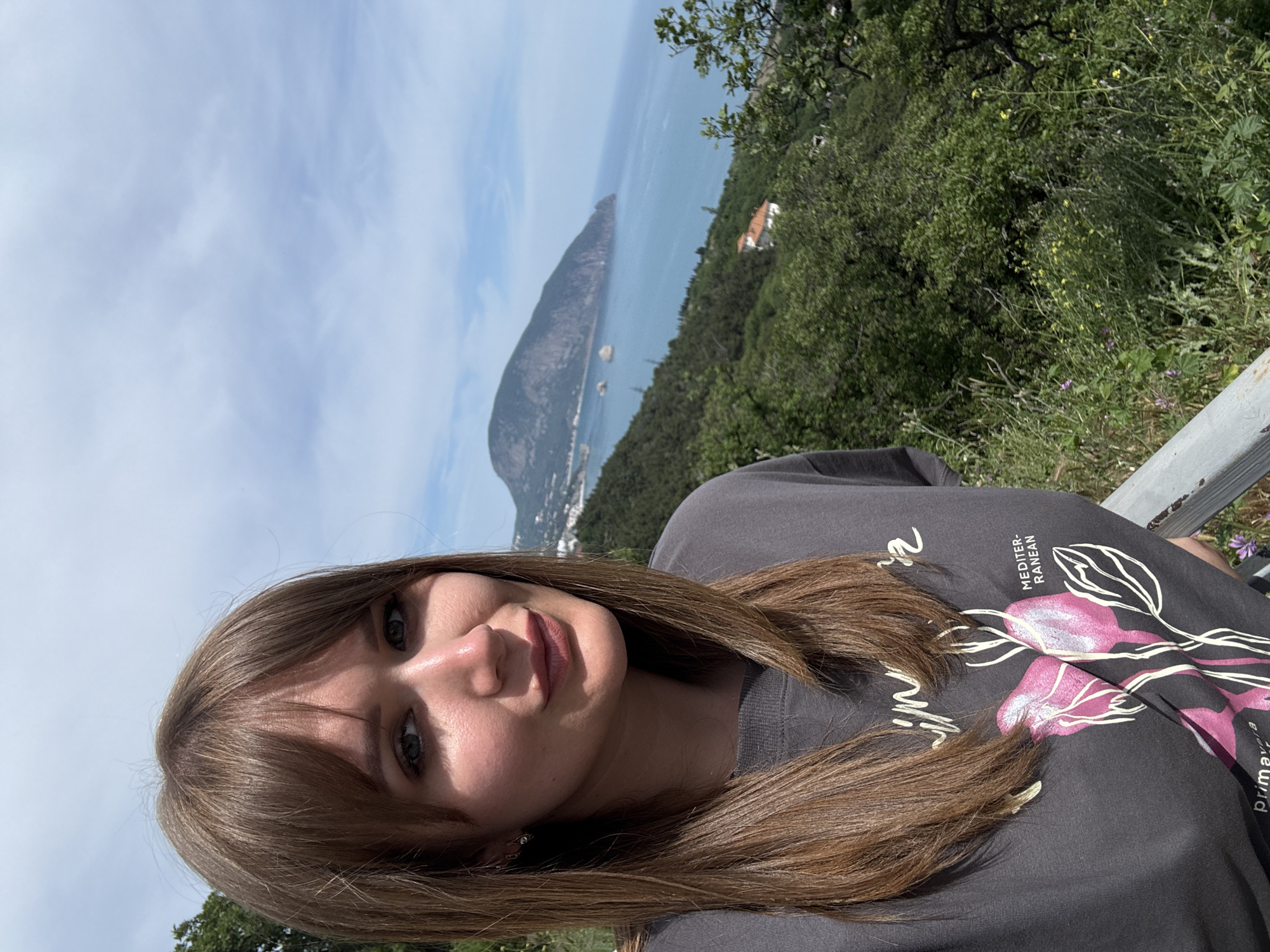 A young woman with long brown hair is pictured from a slightly high angle, likely a selfie. She is wearing a dark gray t-shirt with a floral design. The background includes a mountainous landscape, with a body of water visible in the distance. Some greenery is present, indicating trees and vegetation. In the far distance, signs of habitation can be seen, possibly buildings. The setting appears to be a scenic overlook. The scene is taken in Гурзуфский поселковый совет, Ukraine.