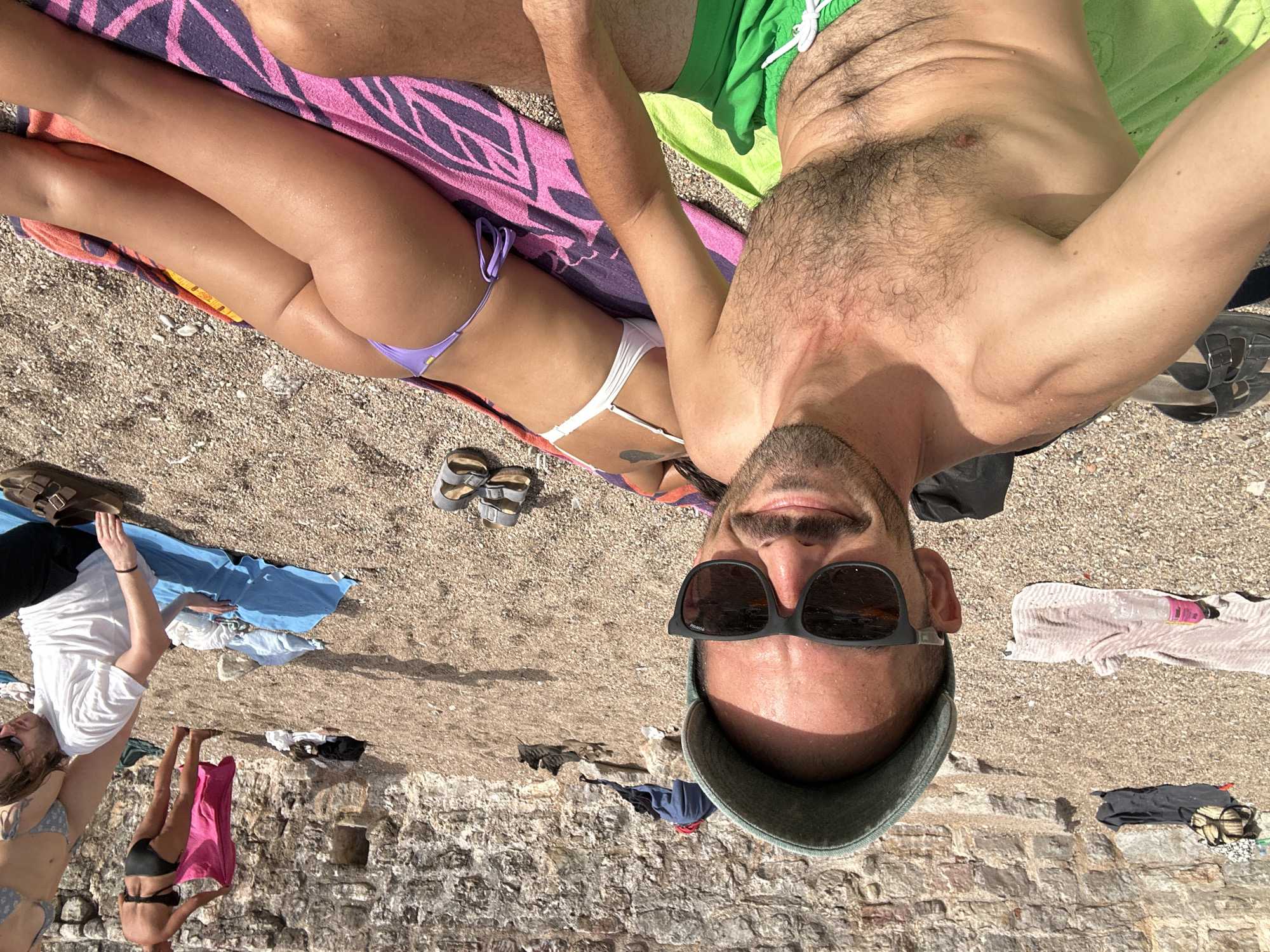In Budva, Montenegro, on a pebble beach with a stone wall backdrop, a man wearing a green cap and sunglasses takes a selfie. He is shirtless, wearing green swim trunks, and has a towel draped over his legs. Next to him, a woman lies on her front on a pink and purple towel, wearing a white bikini top and purple bikini bottoms. In the background, other people are visible on the beach, with towels and clothing scattered around.