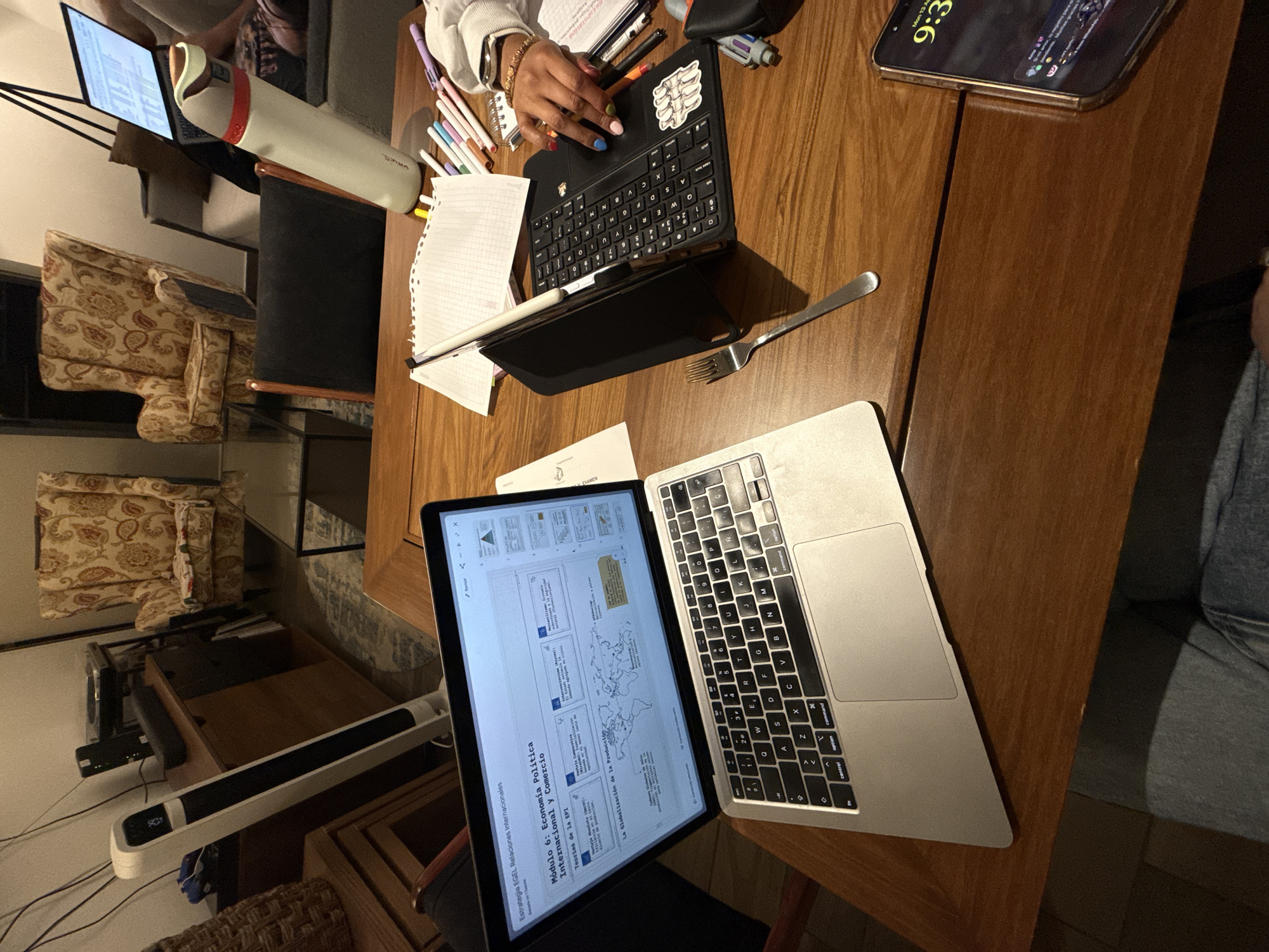 The image captures a scene of study or work in progress, with a wooden table dominating the view. On the table are multiple devices including a MacBook and an iPad, along with a keyboard, a fork, a notebook, and pens. A person with colorful nail polish is using one of the keyboards. A large phone is also visible. The laptop screen displays the words "Módulo 6: Economía Política Internacional y Comercio." In the background, there are two patterned armchairs and a small laptop stand, suggesting a home or study environment. A lamp and other furniture are also in the background. The time on the phone is 9:36. The image doesn't provide information that can indicate it is Mexico City.