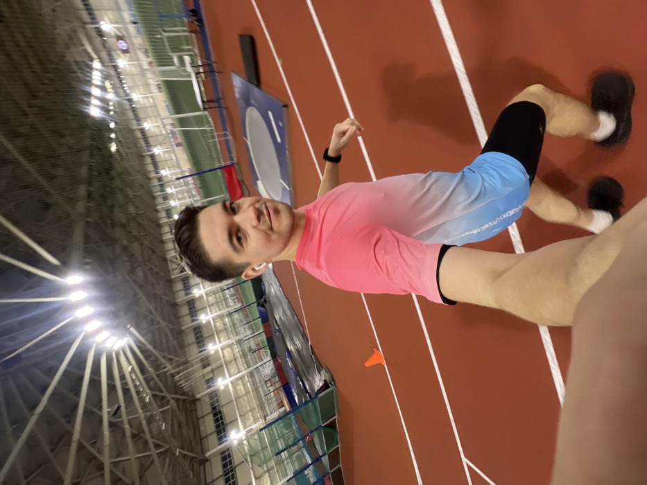 A man stands on a red indoor track, facing the camera. He wears a pink and blue gradient shirt, black shorts, and black shoes with white socks. He has short dark hair, a white earbud in one ear, and a black watch on his left wrist. The track is marked with white lines. A large metal structure is visible in the background, along with stadium lights. The photo appears to be a selfie taken indoors.