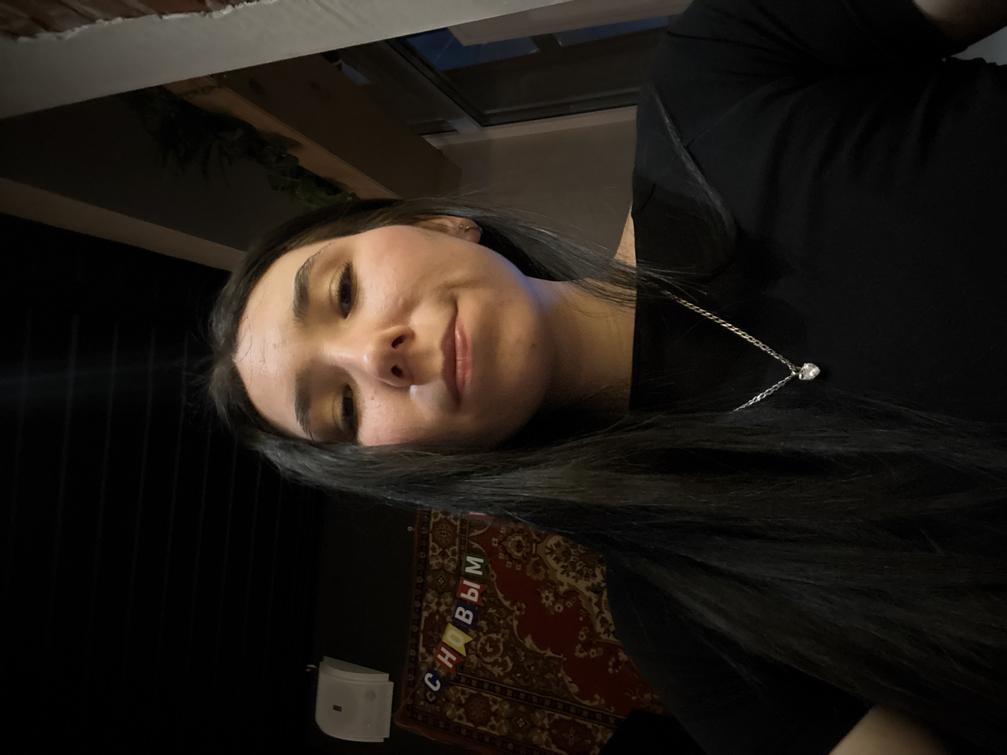 A medium shot captures a person with long, dark hair and light skin, wearing a black top and a silver necklace with a pendant. The person is facing the camera with a slight smile.
Behind the person, a decorative rug with red, brown, and gold patterns and the Cyrillic text "СНОВЫМ" is visible on the wall. To the left of the rug is a small, white, rectangular object mounted on the wall.
The setting appears to be indoors, with dark, vertical lines suggesting blinds or paneling on the left side of the frame. Above, there is a window and part of a ceiling.
While the image provides clues that are consistent with locations found in Izhevsk, Russia, such as the Cyrillic text, the image lacks specific identifying features to definitively confirm this location.