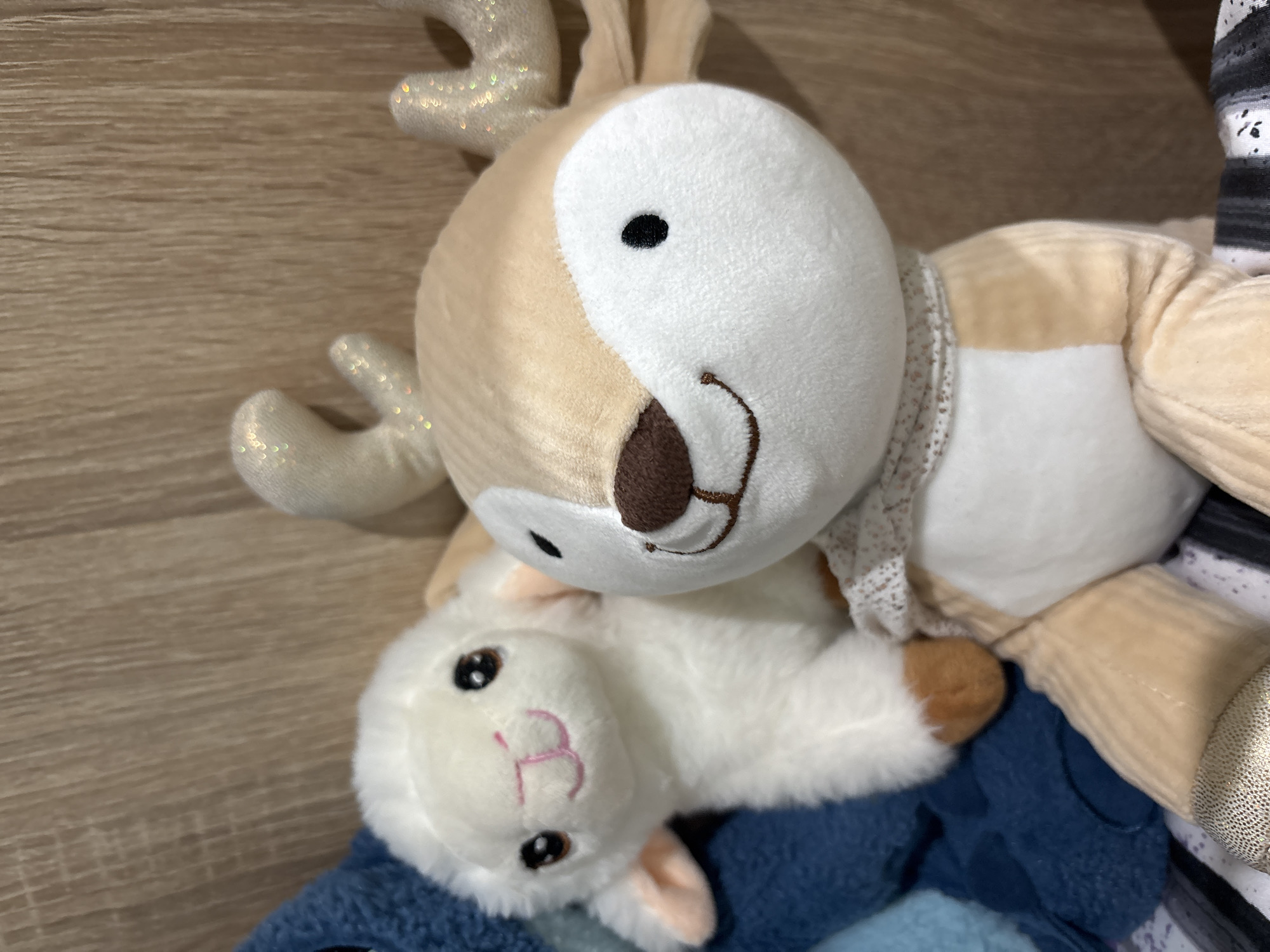 A deer plush toy is stacked on top of a lamb plush toy. The deer plush has a tan body and a white face, while the lamb plush is entirely white. A blue blanket lies beneath the lamb. The background is a wooden surface. The location is indeterminate.