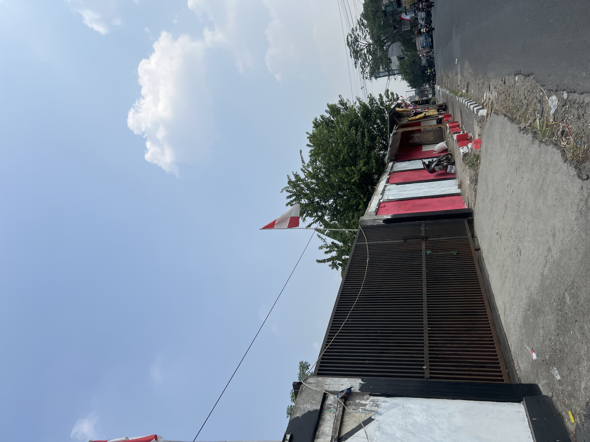 An eye-level, medium shot captures a street scene in Citeureup, Indonesia, under a bright, partly cloudy sky. The foreground features a concrete sidewalk with scattered litter and a dark metal gate with vertical slats. To the right of the gate, a row of buildings with red and white facades lines the sidewalk, with a motorcycle parked in front of one. A slender tree with dense green foliage grows behind the buildings. Beyond, a paved road stretches into the distance, with several vehicles, including motorcycles and cars, visible. Power lines crisscross the upper portion of the frame. Two Indonesian flags, one on a flagpole and another smaller one attached to a building, are prominently displayed. The scene suggests a typical urban environment on a clear day.
