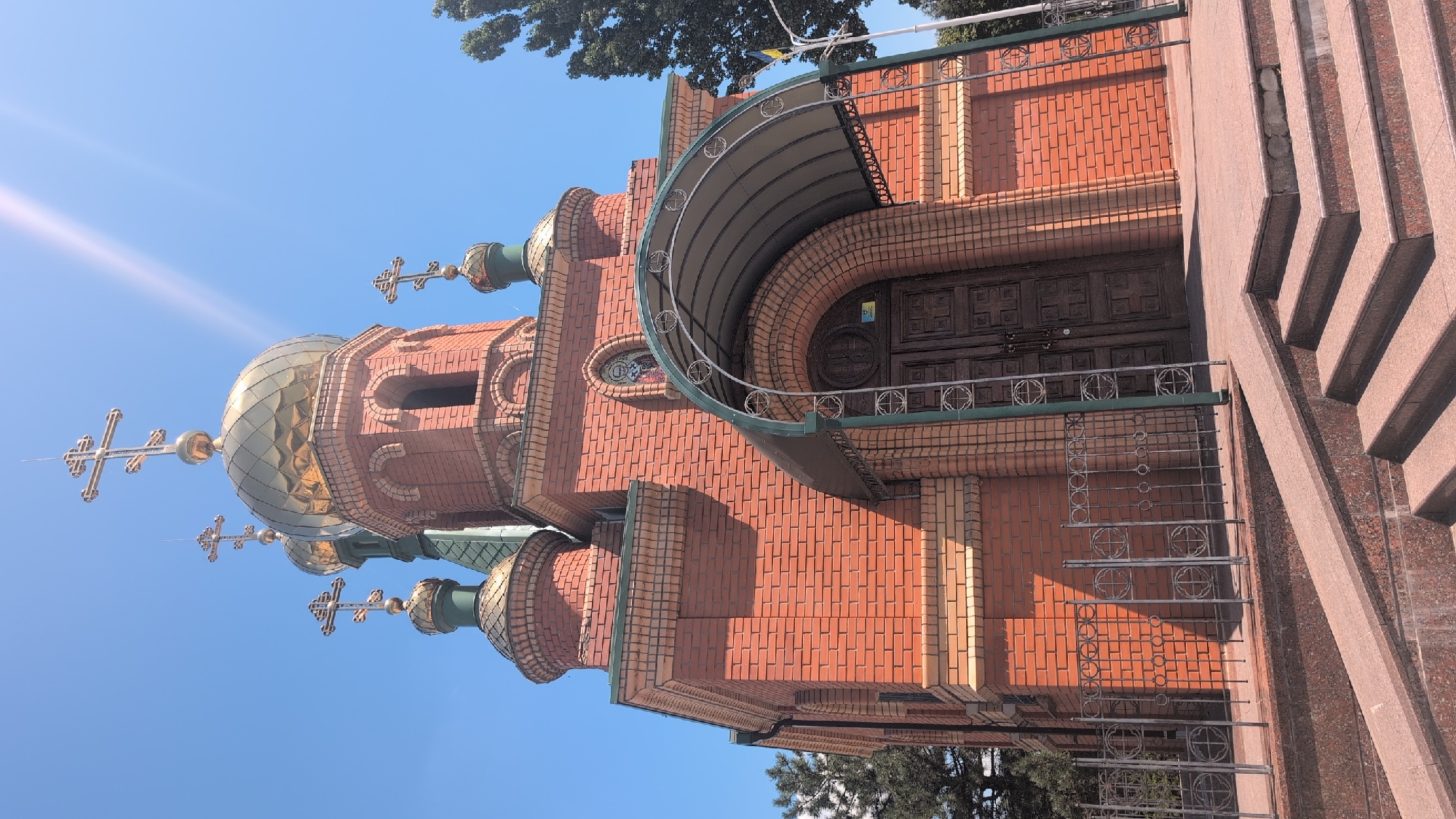 The image shows a close-up, low-angle view of the brick facade of a church, likely the Church of the Intercession of the Theotokos in Izmail, Ukraine, under a clear blue sky. The church features multiple golden domes topped with crosses, characteristic of Orthodox Christian architecture. The main structure is constructed of reddish-brown brick with lighter colored decorative bands. An arched entrance with large wooden doors is partially covered by a dark green metal awning supported by decorative metal arches. A low metal fence runs across the front of the building, and in the foreground, there are granite stairs and a ramp leading up to the entrance. The sun is shining brightly, creating lens flare in the upper right portion of the image and casting shadows on the building and fence. No people are visible in the image.