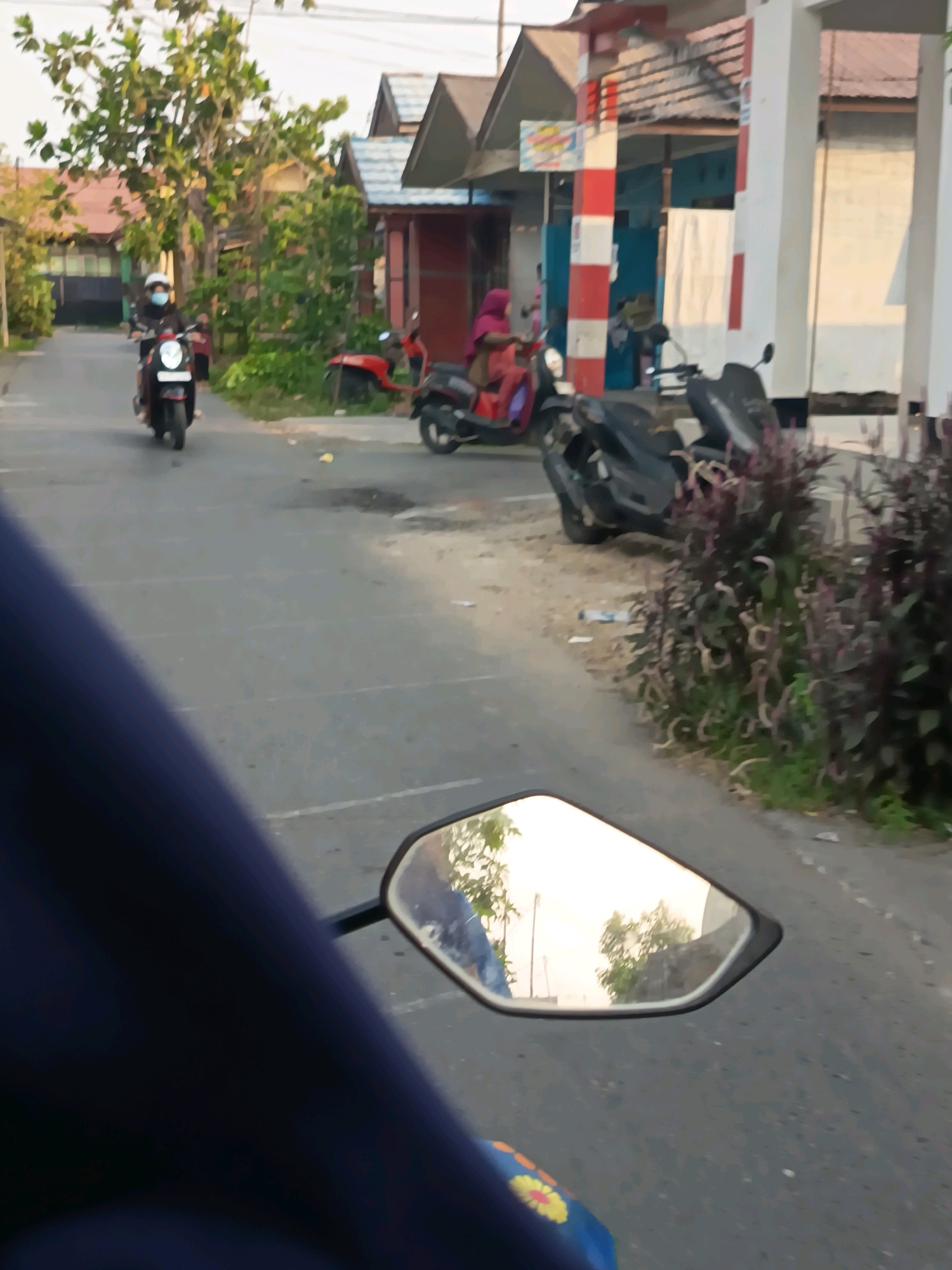 This is a first-person perspective shot from a motorcycle in Banjarmasin, Indonesia, capturing a street scene. A motorcycle with a rider wearing a white helmet and blue mask is visible in the middle ground, moving away from the camera. To the right, another motorcycle is parked, and behind it, a person wearing a pink headscarf is seated on a red scooter. Further down the street, more parked motorcycles are present. The environment includes houses with red tiled roofs and some trees. The road surface appears to be asphalt with some minor cracks and debris. The overall lighting suggests it is daytime.