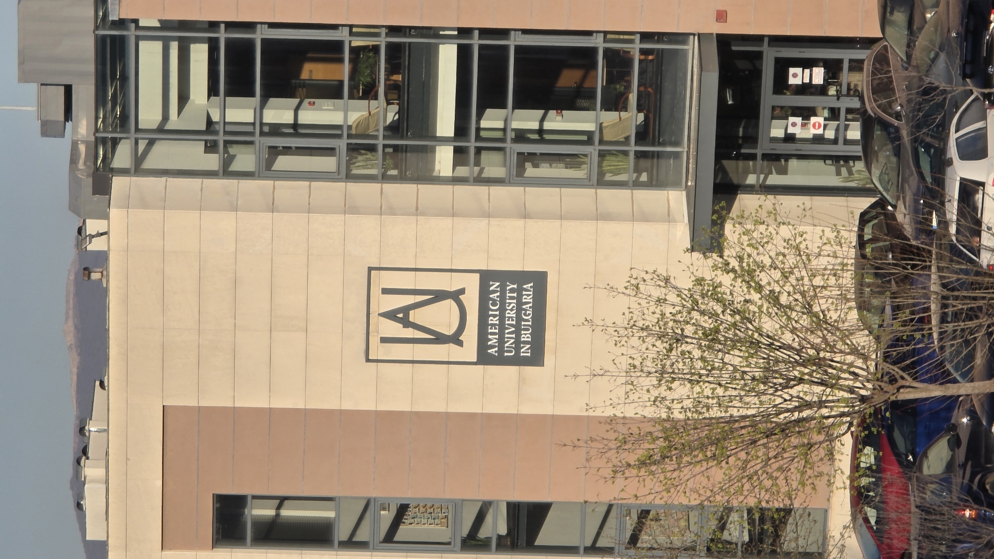 The image shows a building in Blagoevgrad, Bulgaria, identified by a sign as the "American University in Bulgaria". The building has a modern architectural style, with large windows and a light-colored facade. A tree with budding leaves partially obscures the view. Several parked cars are visible in the foreground, suggesting the building is situated in an urban area.