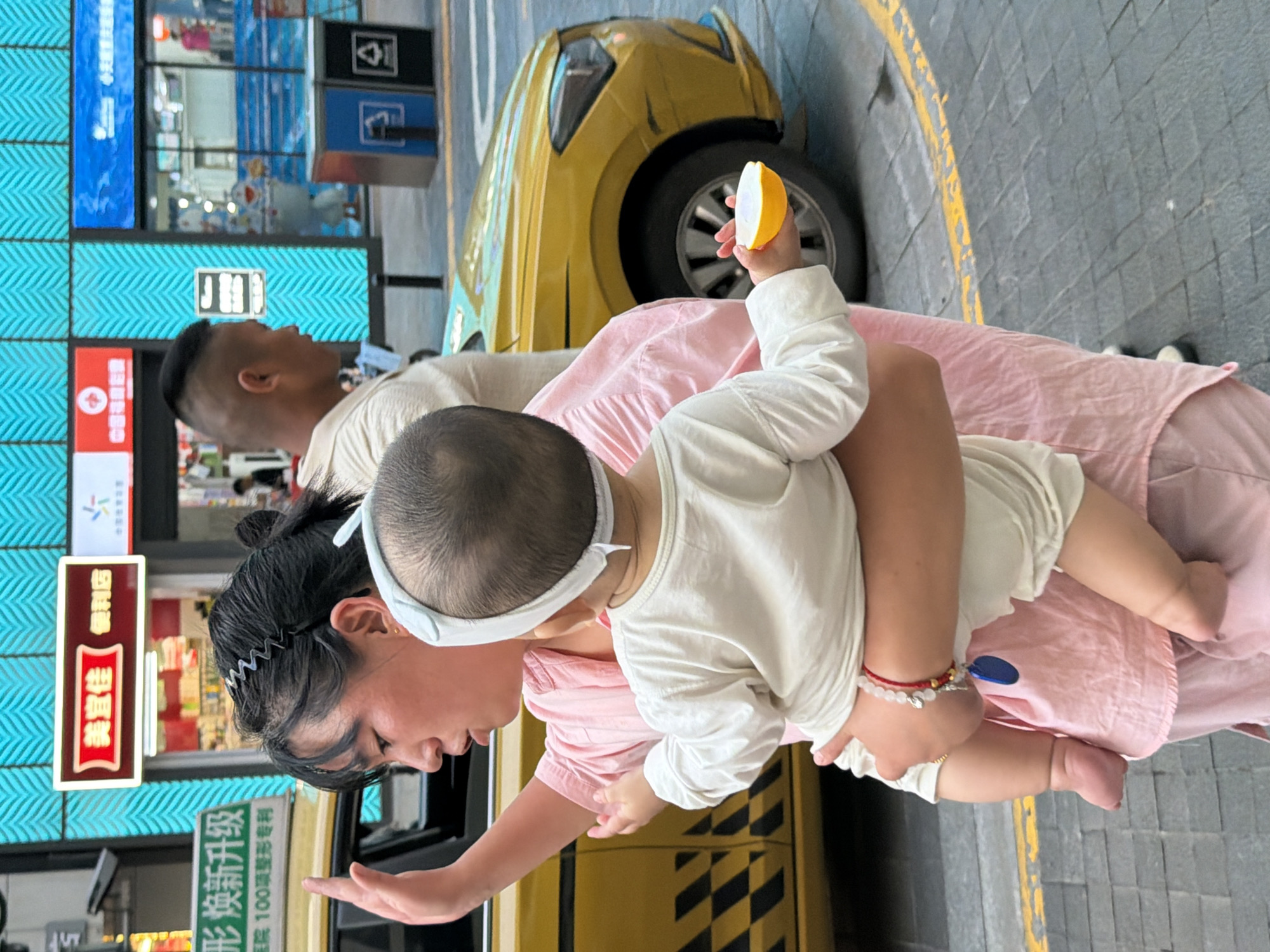 In Yuzhong District, China, a woman in a pink shirt carries a baby. The baby, wearing a white outfit and a white headband, is held in front of the woman. The baby holds a yellow object in their right hand. A yellow taxi is partially visible to the left of the woman, and a man with a shaved head is in the background. The scene takes place on a paved street with yellow and gray markings. Behind the individuals, a building facade features bright blue and red signage. Two trash receptacles, one blue and one black, are situated on the right side of the frame. The lighting suggests daytime.