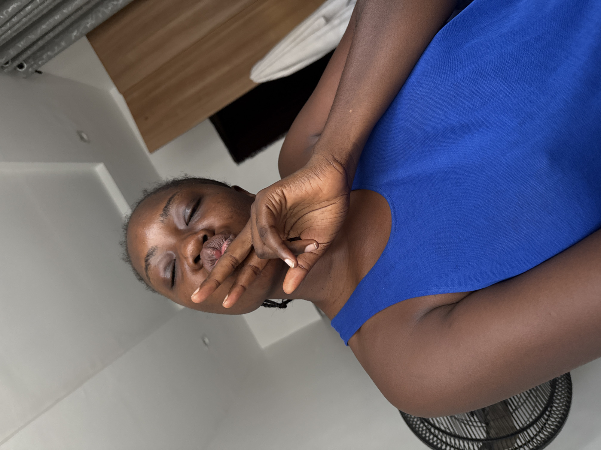 A dark-skinned woman wearing a blue top is shown with her eyes closed and her fingers near her mouth. A fan sits nearby. The setting is a room with light-colored walls and ceiling. The image location is Iddo Tudunwada, Nigeria.