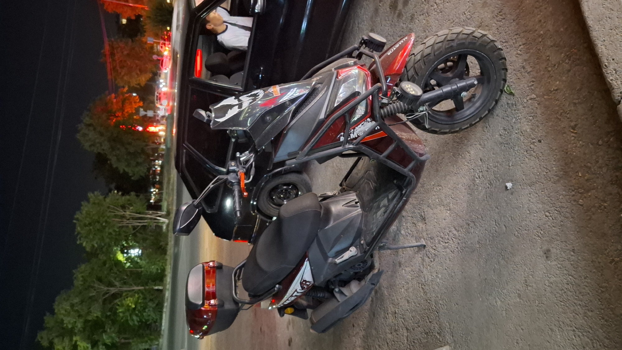 A red scooter is parked on a gravelly surface next to a black car at night. The scooter has a black seat, a red top box, and a black windshield with a skull graphic. The car has its trunk open, and a person wearing a white shirt is visible in the driver's seat. Streetlights and trees are in the background, with some red and orange lights indicating traffic or a business. The location is the city of Shymkent, Kazakhstan.