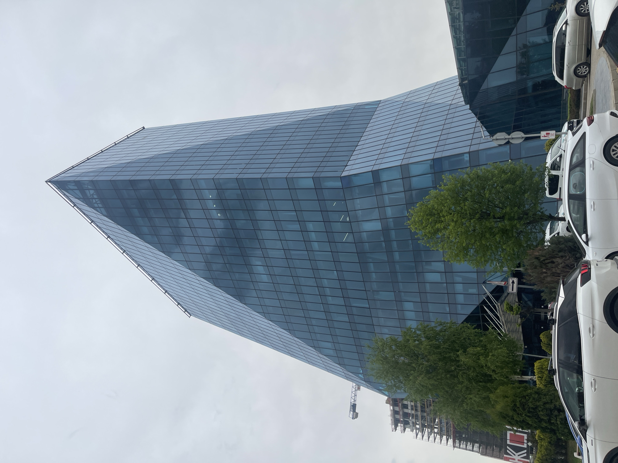 The image features a modern, glass-clad skyscraper in Sofia, Bulgaria. The building has a unique geometric design, tapering to a point at its top. Its glass facade reflects the cloudy sky above. At the base of the building, there are trees and several parked white cars. In the background, partially obscured by trees, there is construction work with a crane. The overall environment suggests a business or commercial district within the city.