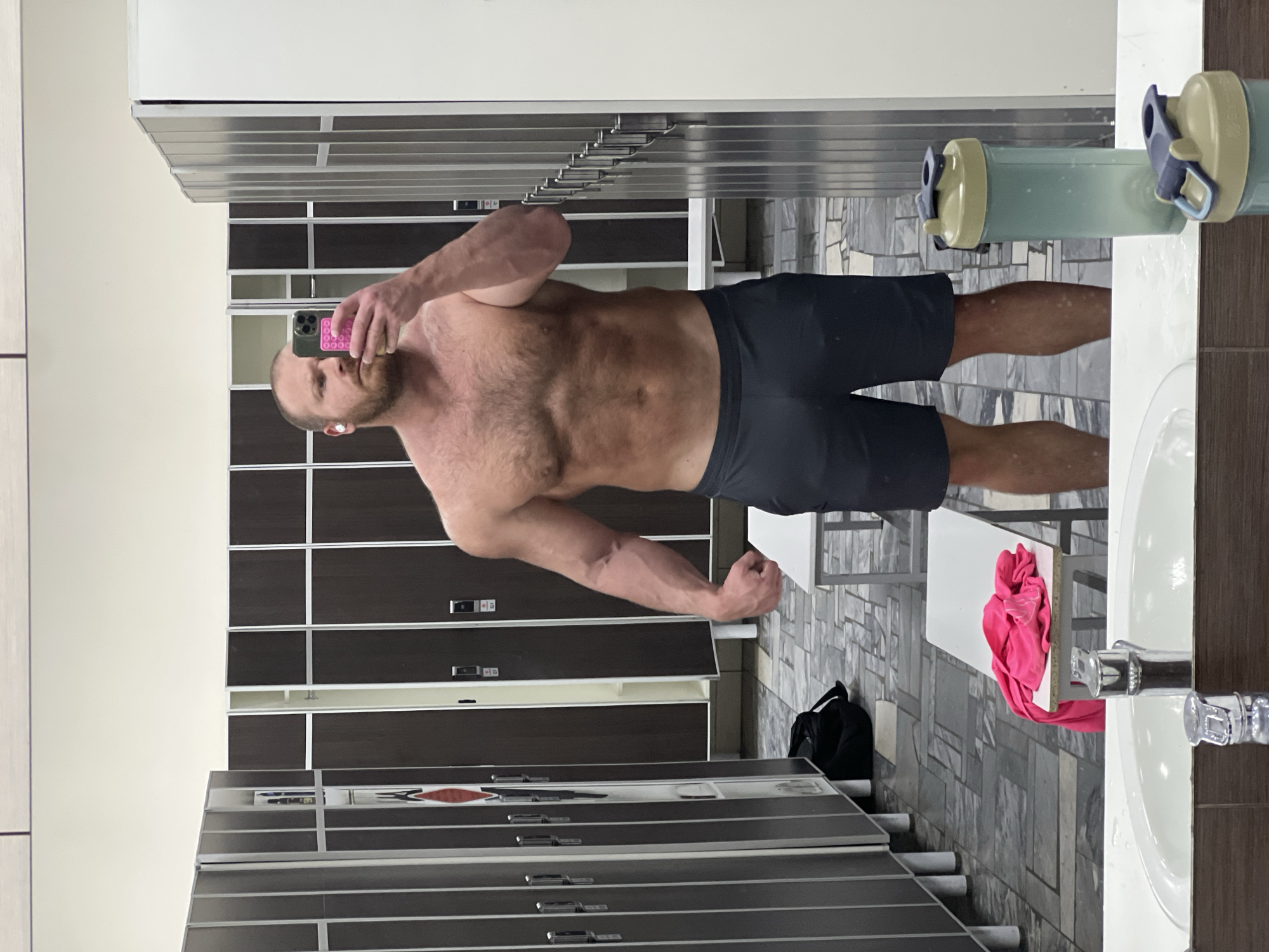 The scene is a gym changing room, located within the city of Chaikovsky, Russia. A shirtless, muscular male subject with a beard and short hair stands centered in a mirror, taking a selfie with a smartphone held in his right hand. He wears dark grey athletic shorts and a white earbud in his left ear. The smartphone has a distinct pink, textured protective case. His right arm is slightly flexed.
The changing room features light-colored walls and a floor covered with grey, white, and black tiles arranged in geometric patterns. Along the left and background, numerous individual lockers with dark brown or black doors and light grey or white vertical frames are visible. Some locker doors are ajar, revealing light-colored interiors.
In the mid-ground, two white, rectangular benches are present. A bright pink piece of fabric, possibly a shirt or towel, is folded and resting on the bench situated near the center-left. A black duffel bag is on the tiled floor near the base of the lockers on the left.
In the immediate foreground, a white countertop with a built-in sink and a chrome faucet is visible. To the right of the sink, a light green and beige shaker bottle stands upright on the counter, partially filled with liquid, and a portion of a second shaker bottle or its lid is visible in the reflection closer to the bottom right.