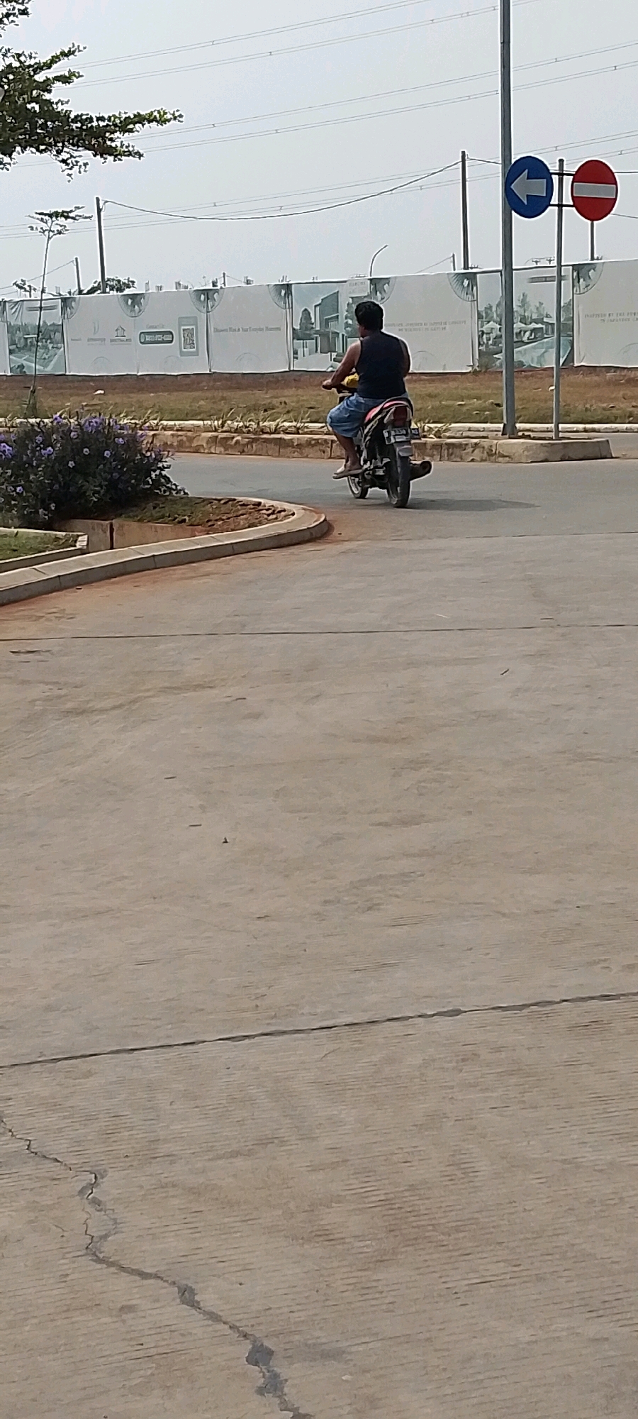 In Satriamekar, Indonesia, a man rides a motorcycle on a paved road. He is wearing a dark tank top and light blue shorts. The motorcycle is black with red and white accents. The road curves to the left in the foreground. In the background, a construction site is visible with a large white tarp fence displaying images of houses. Traffic signs, a blue arrow pointing left and a red circular sign with a horizontal white bar, are mounted on a pole to the right of the motorcycle. Overhead power lines are present. The sky is overcast.