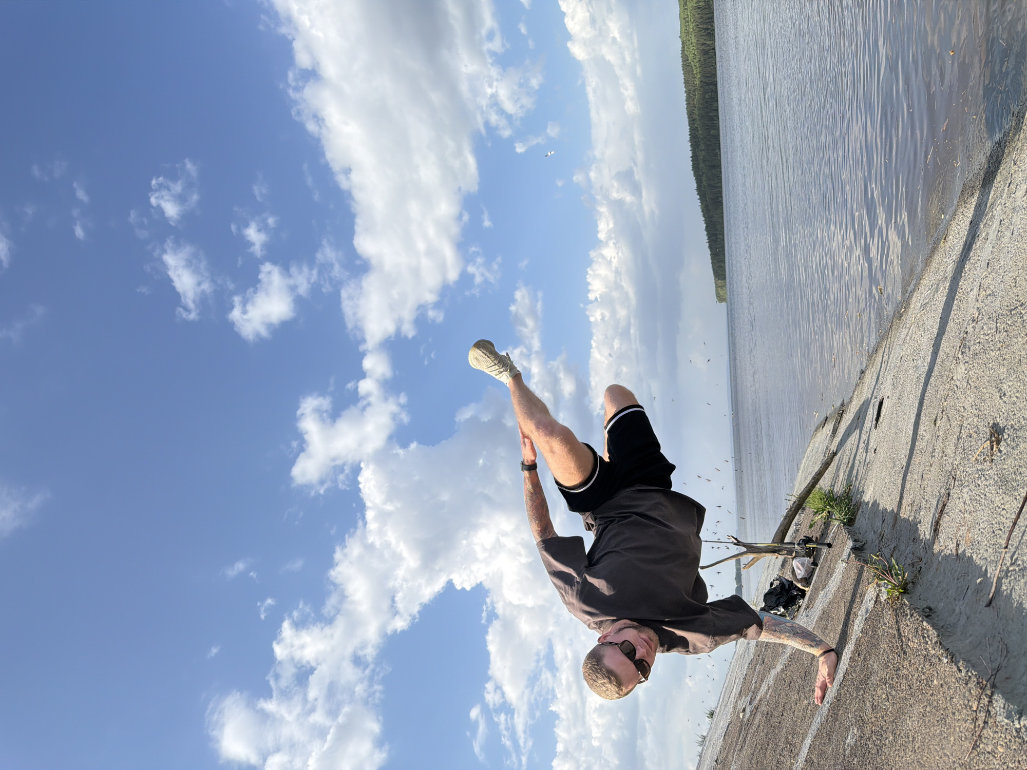 A man performs a handstand on a concrete embankment adjacent to a body of water, likely a lake or reservoir. The sky above is blue with scattered white clouds. The man is wearing shorts and a t-shirt and has visible tattoos. His feet are pointed towards the sky. A stick and some vegetation are visible on the embankment. The photo was taken in the city of Chaikovsky, Russia.