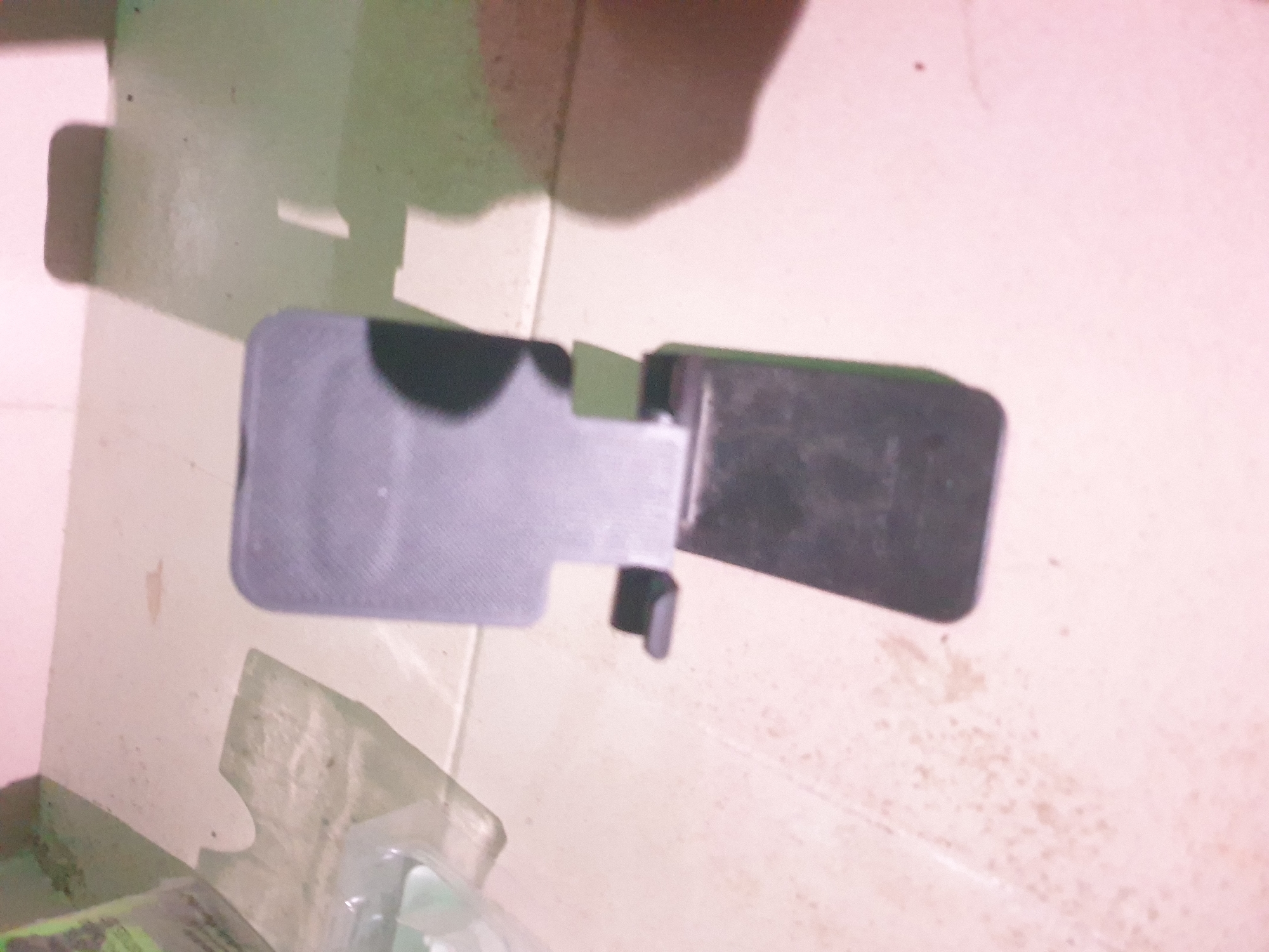 A black plastic object, resembling a phone stand, is positioned on a light-colored, possibly tiled surface. The object appears to be made of two main parts connected by a hinge. The upper part is a flat rectangle with a textured surface, and it is propped up by a smaller, rectangular base. The lighting in the scene creates distinct shadows on the surface, suggesting an overhead light source. Some indistinct objects and shadows are visible in the background, including what might be a plastic bag on the left side. The overall environment appears to be indoors, possibly a room or a sheltered outdoor area, with the floor showing some minor dirt or dust. The location within Anilelerin, Nigeria, cannot be determined from the visual information provided.