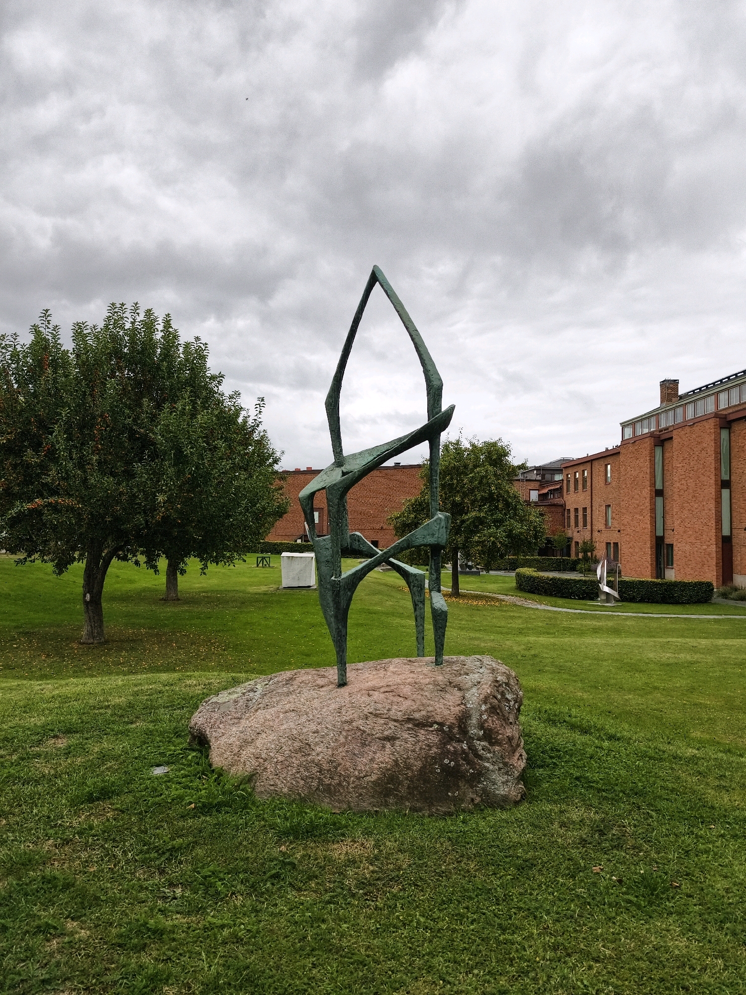 A bronze sculpture of an abstract figure stands on a large rock in a grassy park. The sculpture is dark green and appears to be in motion. Behind the sculpture, there are trees and a brick building. The sky is overcast. The sculpture is located in the Courtyard of the Industrial Museum in Norrköping, Sweden.