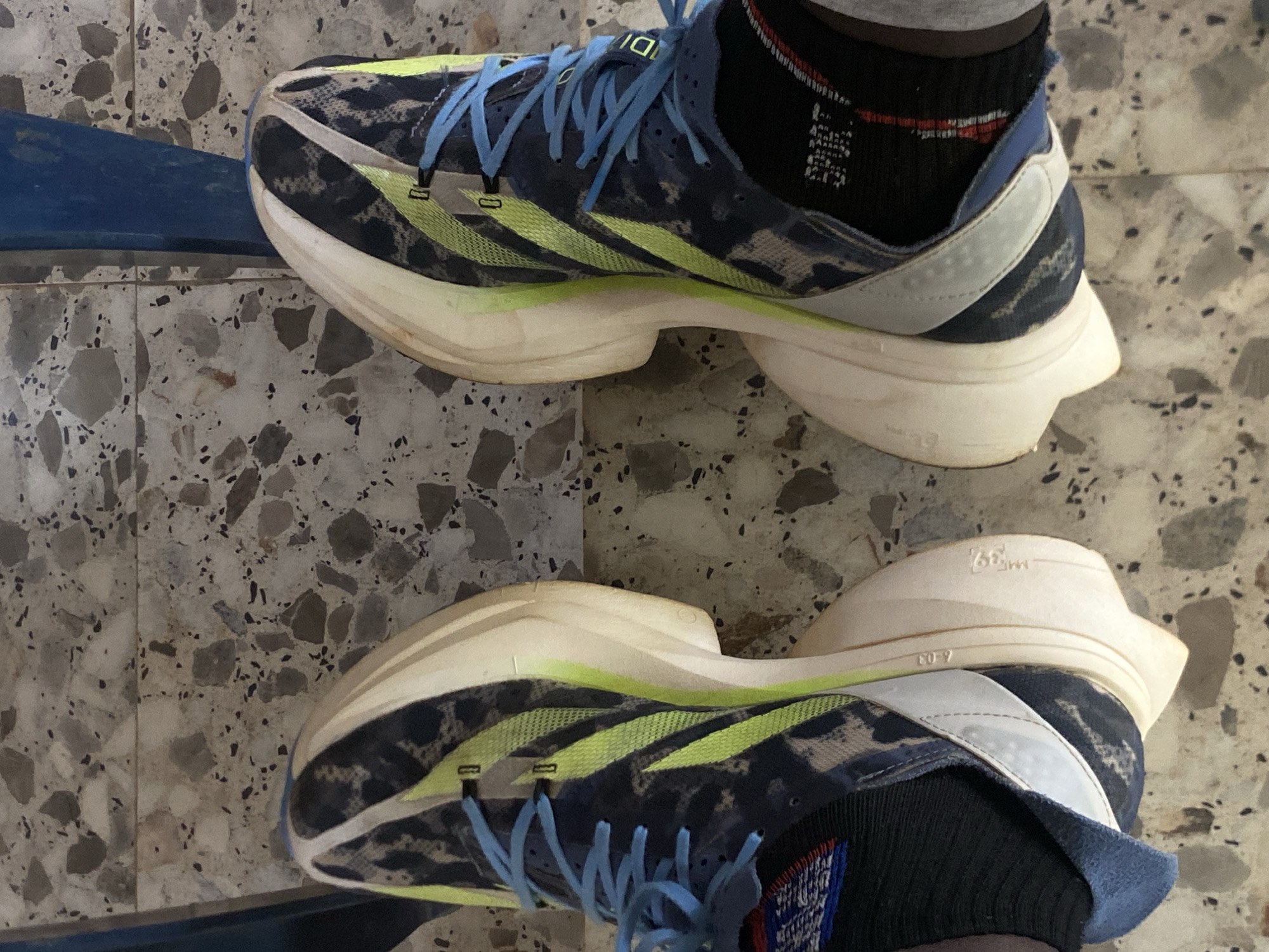 The image shows a pair of Adidas Adizero Adios Pro 3 running shoes on a patterned tile floor. The shoes are blue and black with neon yellow stripes and blue laces. The soles are white and show some wear. The person wearing the shoes has black socks on with a red and blue pattern. The background is out of focus, but the patterned floor suggests an indoor setting. There is a hint of a blue object in the upper left corner. The image doesn't contain enough information to determine if it was taken in Barden Yana, Nigeria.
