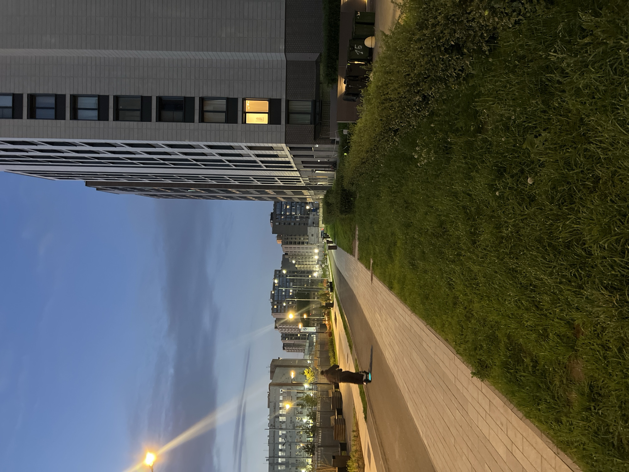 The image depicts an urban street scene at twilight or early night in Saint Petersburg, Russia. A paved asphalt path occupies the center-left, bordered by a light-colored rectangular paver walkway to its right. A single individual, wearing dark clothing, is riding an electric scooter with a prominent blue underglow, moving away from the viewer along the asphalt path.
On the right side of the image, a strip of unkempt green grass with interspersed wildflowers runs parallel to the paver walkway. Beyond this grass and a hedge of dark green bushes, a modern, multi-story residential building with a light grey panel facade and uniformly spaced dark-framed windows dominates the midground. One window on the lower-mid section of this building emits a warm yellow light. At the base of the building, a cluster of dark green waste bins is visible, with the number "15" marked in white on one.
On the left side of the scene, across the asphalt path, multiple streetlights with bright yellowish-orange illumination cast light onto another paved walkway and the surrounding areas. These lights generate noticeable lens flares, particularly in the upper left of the image. In the background on the left, an older, multi-story grey building with numerous windows and visible utilities stands behind a metal fence. Further beyond, additional multi-story residential structures are discernible. The sky above is a deep blue, transitioning to a lighter blue towards the horizon, with scattered grey clouds.