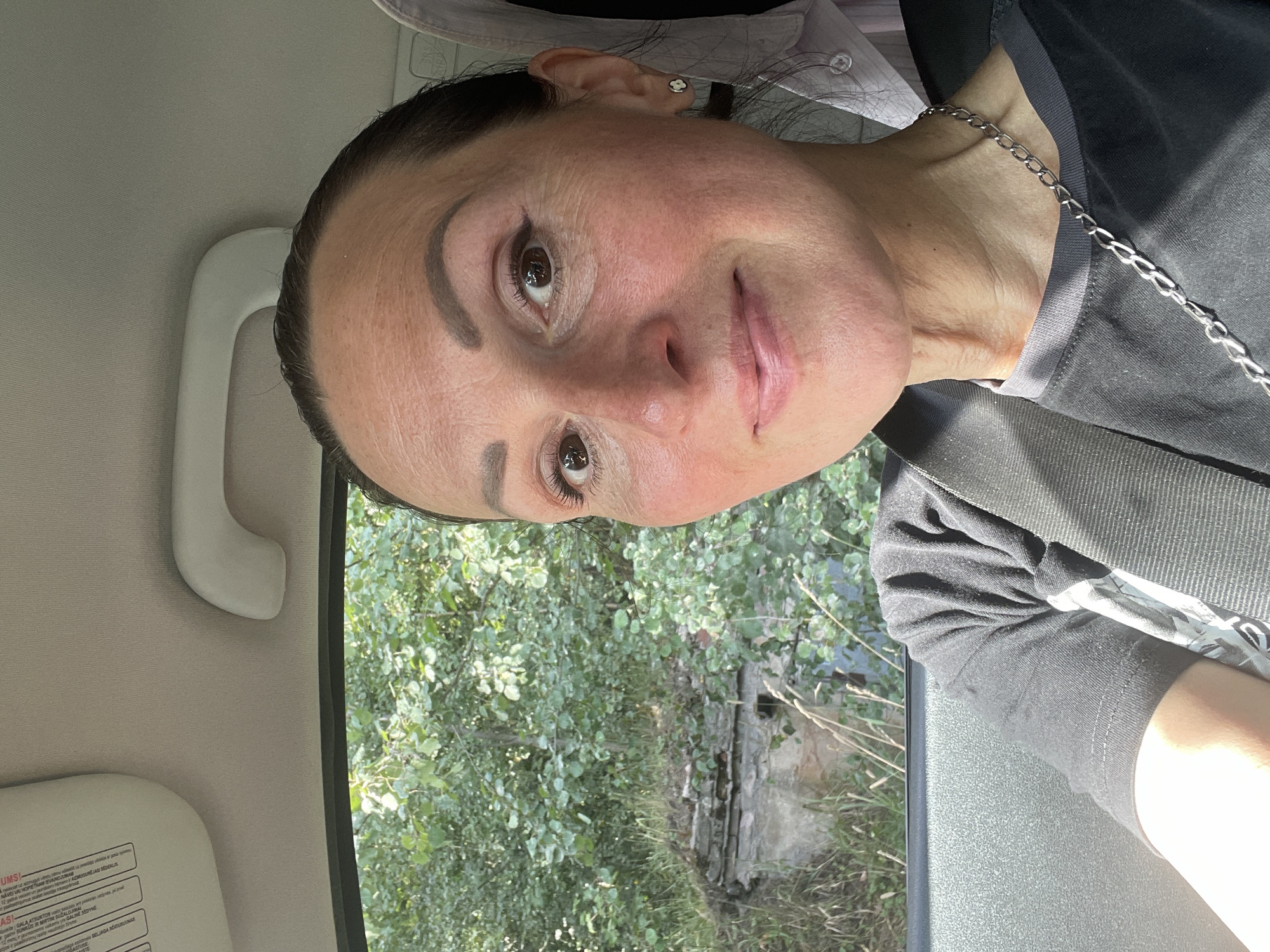 A woman is seen from the chest up, inside a vehicle. She is looking directly at the camera. She has dark hair pulled back, is wearing a black t-shirt, and a silver chain necklace. The background shows a window with trees and what appears to be an old, crumbling structure. The location is stated to be Berozovo, Russia.