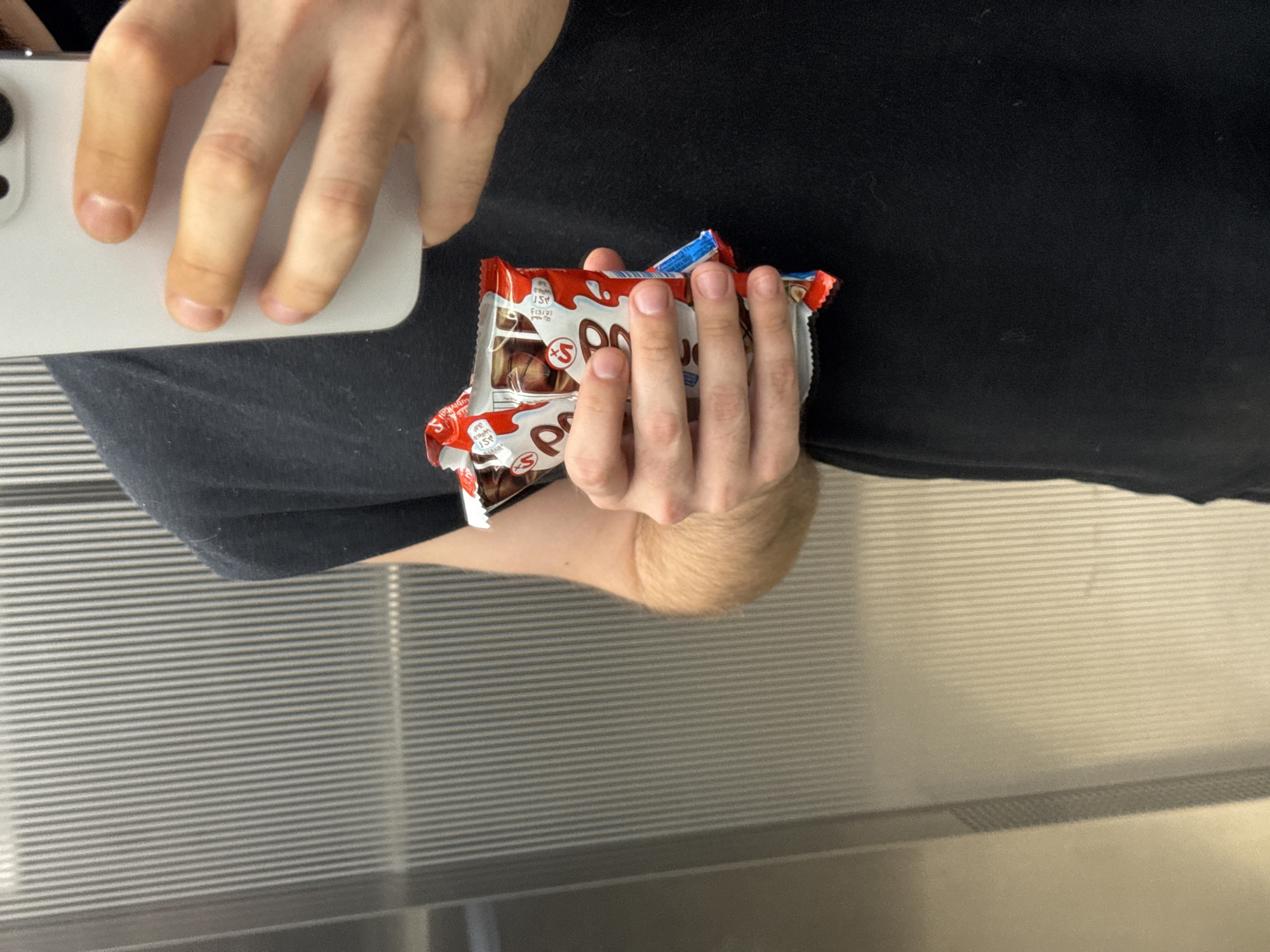 A close-up shot shows a person wearing a black t-shirt holding a Kinder Bueno chocolate bar and a white smartphone in their hands. The background is a metallic ribbed wall, possibly an elevator or escalator. The person's hands are visible, one holding the chocolate and the other holding the phone, suggesting they might be taking a selfie. The lighting is somewhat dim, creating a focused atmosphere on the subject and the objects. The scene is devoid of any recognizable landmarks or context that would indicate its location within Dubai, United Arab Emirates.