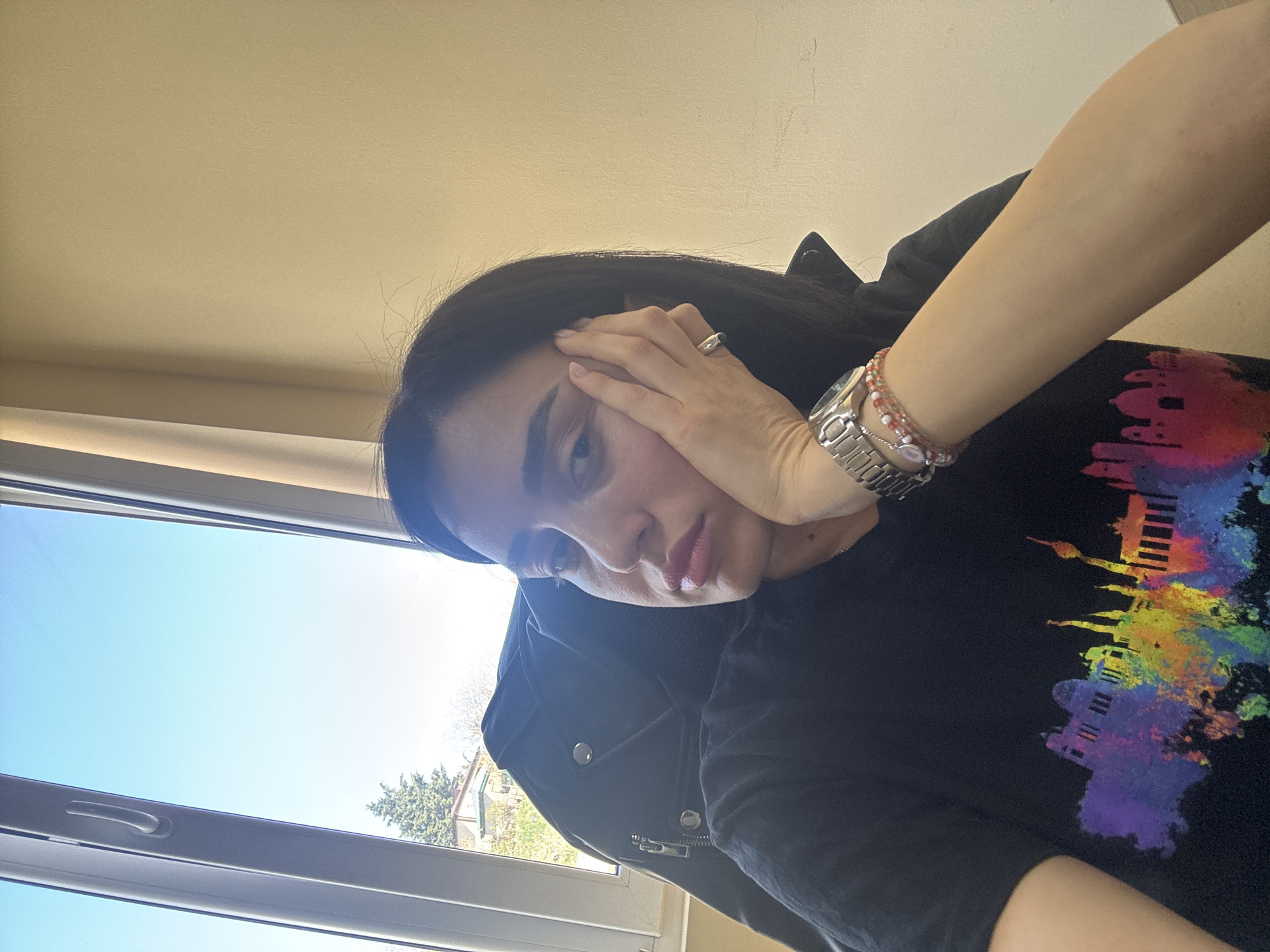 A woman is posing indoors near a window with a view of a building and trees outside under a blue sky. The woman has dark hair and is wearing a black t-shirt with a colorful skyline graphic. She is also wearing a black jacket, a silver wristwatch, and a beaded bracelet. She is touching her face with her hand, which has a ring on one of the fingers. The background suggests an interior setting with a window and a view of the outside. Given the skyline graphic on her shirt, she might be located in Tbilisi, Georgia.