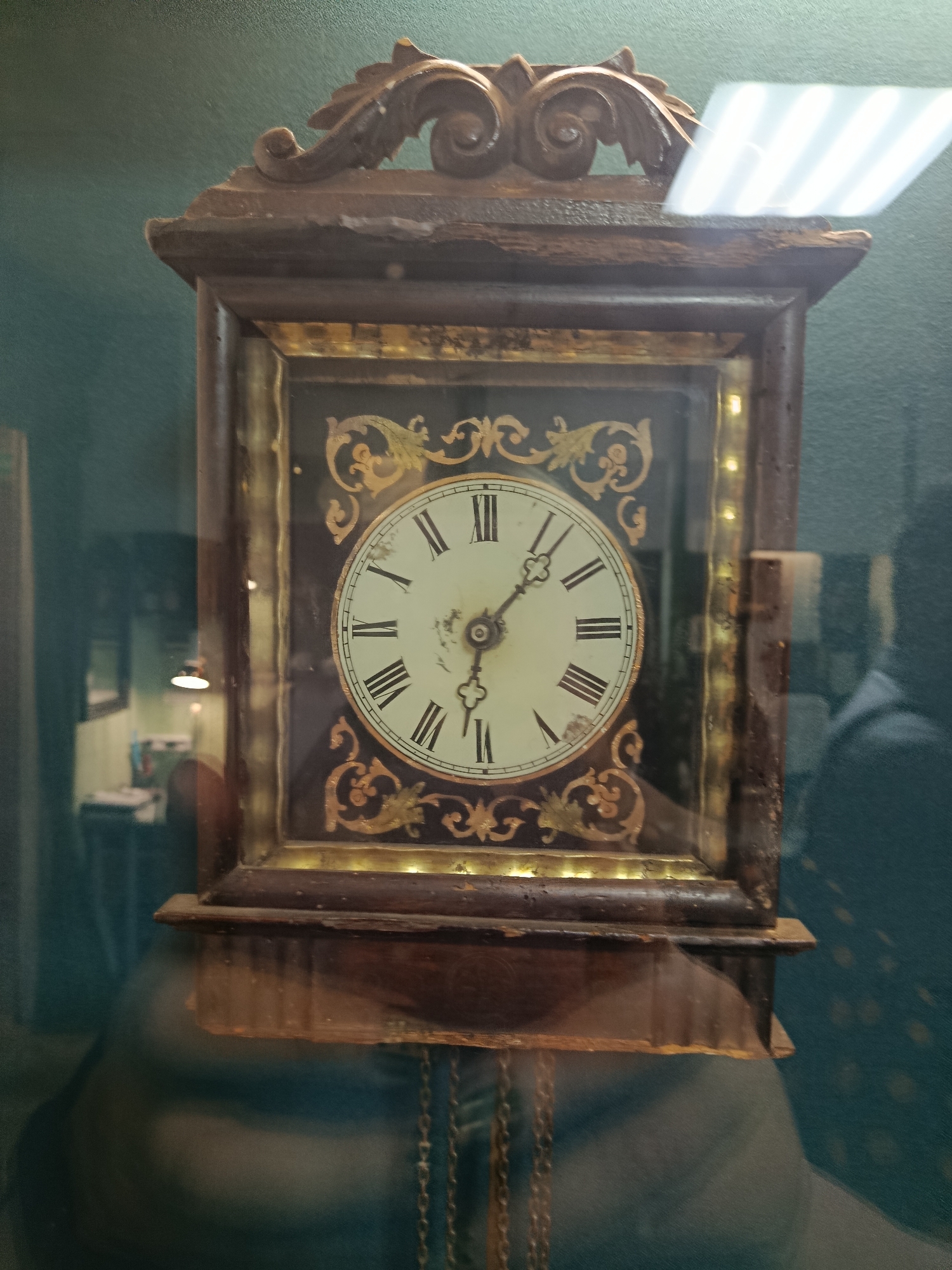An antique, wooden grandfather clock with a glass face is displayed against a dark green, textured wall. The clock face has Roman numerals, and a single hand is visible, pointing to approximately 10:10. Decorative gold filigree adorns the face. The clock is ornately carved at the top and has visible chains hanging from its base. Reflections on the glass suggest the presence of overhead lighting and possibly other objects or people in the room, including a blurred silhouette of a person to the right. The overall setting appears to be a museum or display case. The location of this clock is not specified in the image or provided context.