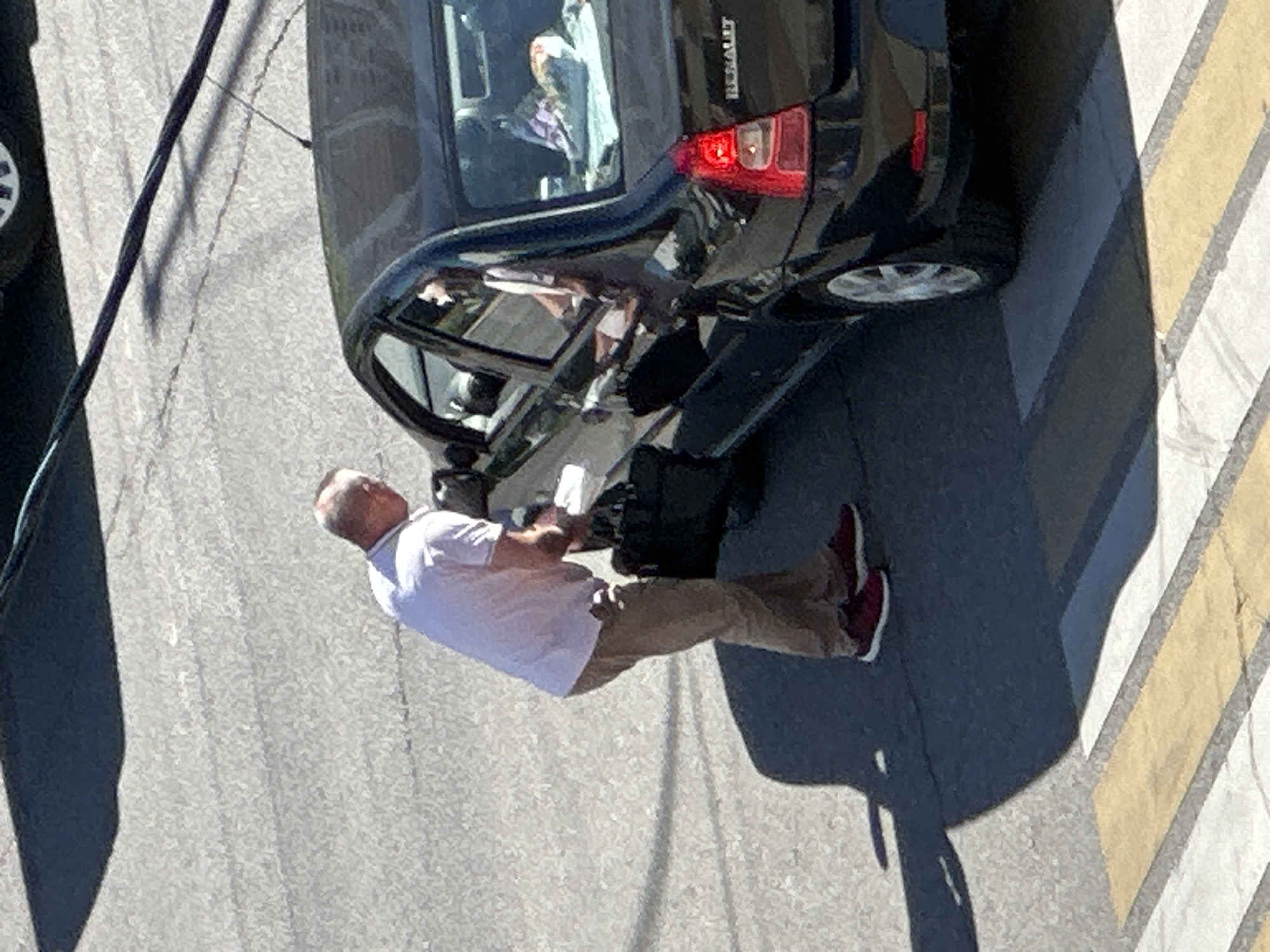 A man in a light pink t-shirt and khaki pants is standing next to an open car door, leaning in to interact with someone inside the vehicle. He is wearing red sneakers. The black sedan has its driver's side door open and the window down. The interior of the car is visible, with items on the back seat. The scene appears to be on a paved road, possibly near a crosswalk, indicated by the yellow and white stripes in the foreground. The lighting suggests it is daytime, with a shadow cast by the man and the car.