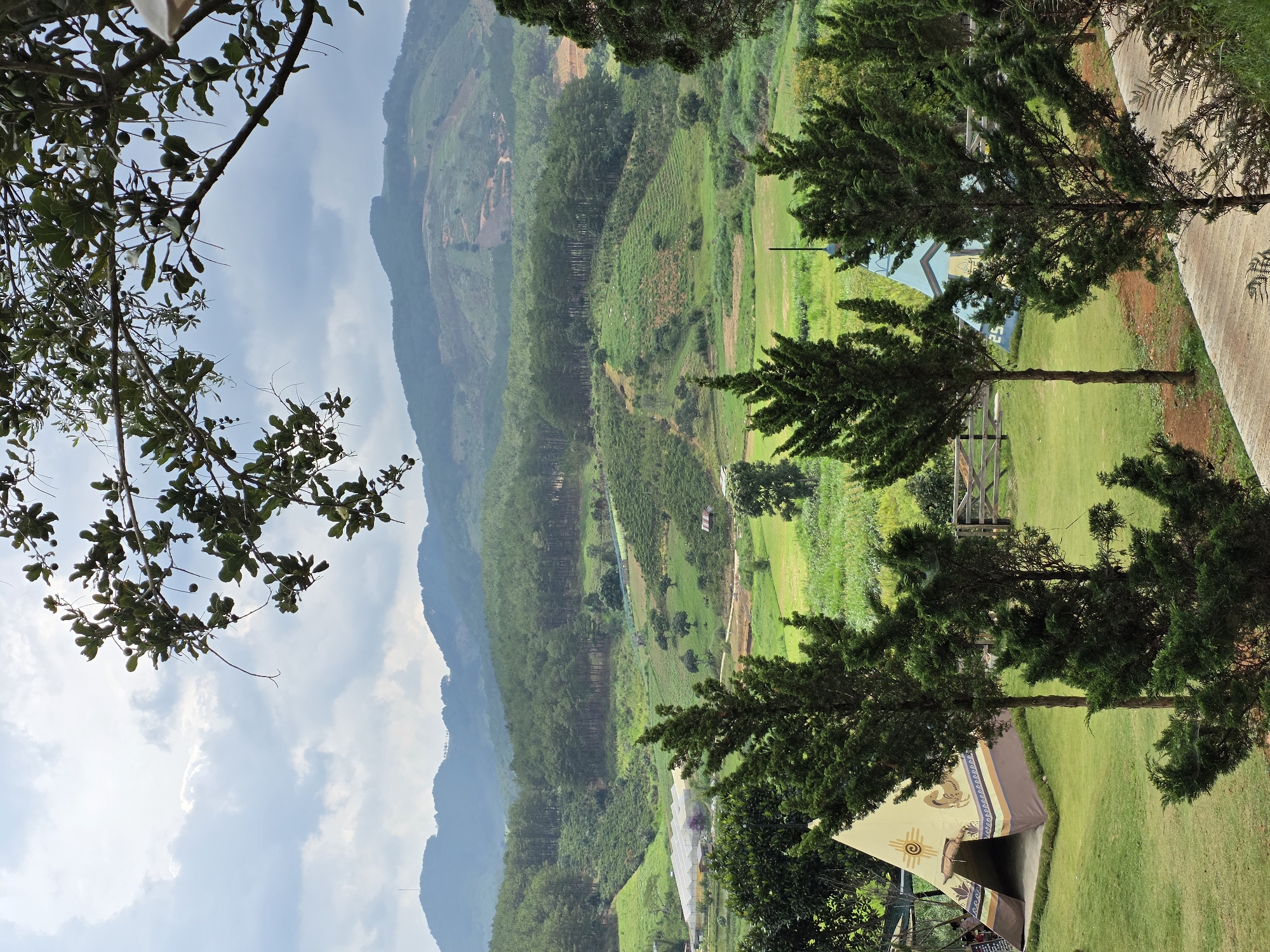 The image depicts a mountainous landscape in Thôn 5, Vietnam. In the foreground, there are trees and some structures, including a teepee-style tent. A paved pathway runs along the right side of the frame. In the midground, a vast, green valley with scattered vegetation and possible cultivated fields stretches towards a mountain range in the background. The sky is partly cloudy. The overall environment appears to be rural, scenic, and hilly.