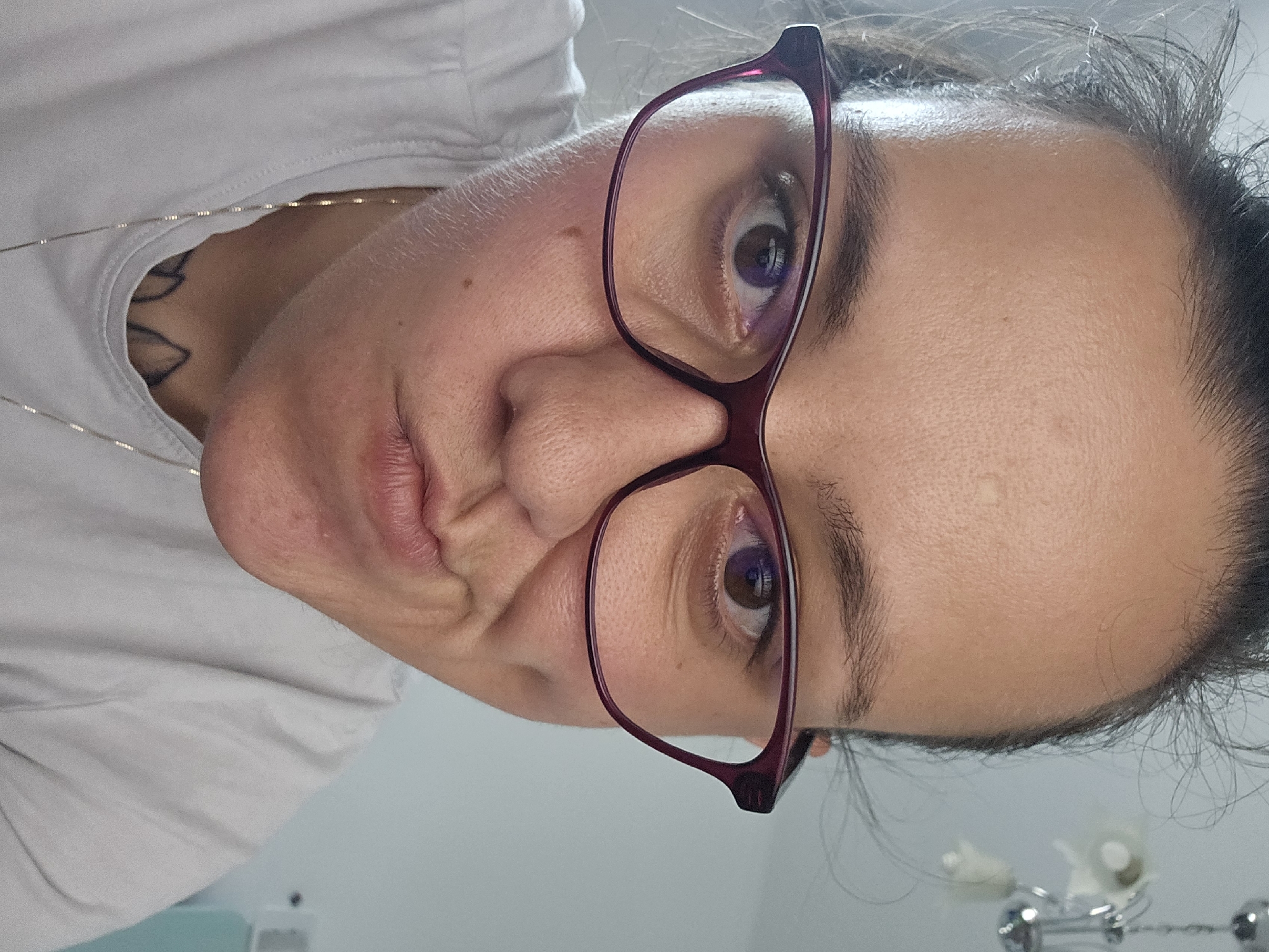 This is a close-up, waist-up photograph of a person wearing glasses. The person is looking directly at the camera with their lips puckered. They have dark hair pulled back and are wearing a light-colored t-shirt, with a silver necklace visible. The background appears to be an indoor setting with a light-colored wall and a chandelier. There is no visible indication of the city of Sofia, Bulgaria.