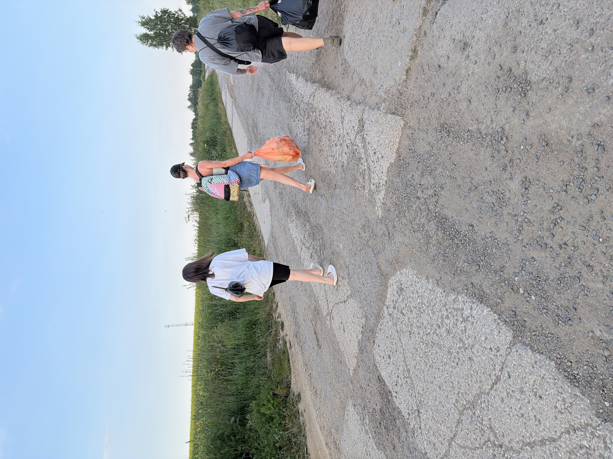 The image shows three people walking on a dirt road, bordered by a grassy field and a clear blue sky. The time of day appears to be late afternoon or early evening due to the soft lighting and long shadows.
The person in the foreground is a woman wearing a white t-shirt and black shorts, carrying a black bag slung across her chest. She is walking away from the camera, her feet clad in white shoes.
Behind her, another woman is walking, also away from the camera. She is wearing a denim skirt and a pink top, and she is carrying a colorful backpack. In her right hand, she holds a bright orange plastic bag filled with objects, possibly groceries or trash. She is wearing white sandals.
Further up the road, a third person, who appears to be a man, is walking towards the camera. He is wearing a grey t-shirt, black shorts, and dark shoes. He has a black backpack on his left shoulder and is carrying a black trash bag in his right hand. His arms are covered in tattoos.
The road itself is unpaved and appears to be made of gravel and dirt, with some larger stones and concrete patches. The field beside the road is lush and green, with some tall grass and scattered wildflowers. In the distance, on the horizon, are a few trees and what looks like a telecommunications tower. The overall atmosphere is casual, suggesting a leisurely walk or perhaps a journey. No text is visible in the image.